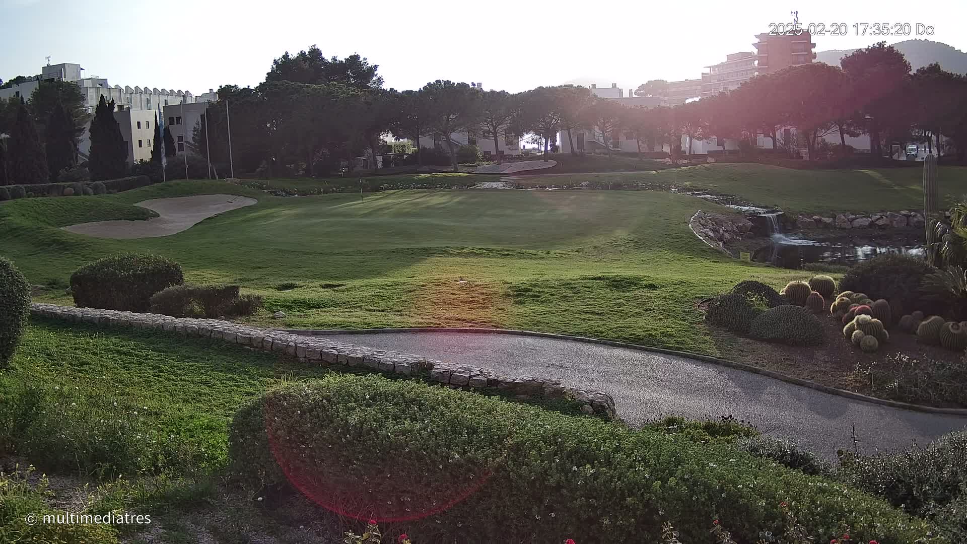 A sunny, partly-shadowed golf course with a sand trap, a small pond with a waterfall, and various shrubs and cacti.
