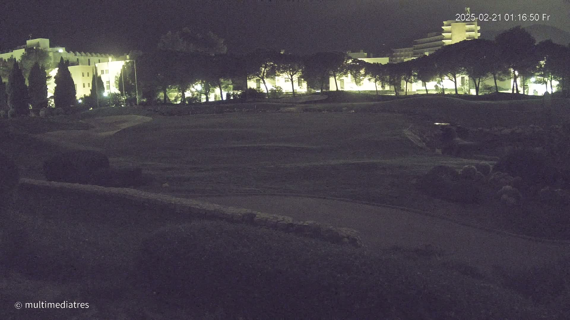 A nighttime, low-light view of a grassy field with a low stone wall and a line of illuminated buildings and trees in the background.