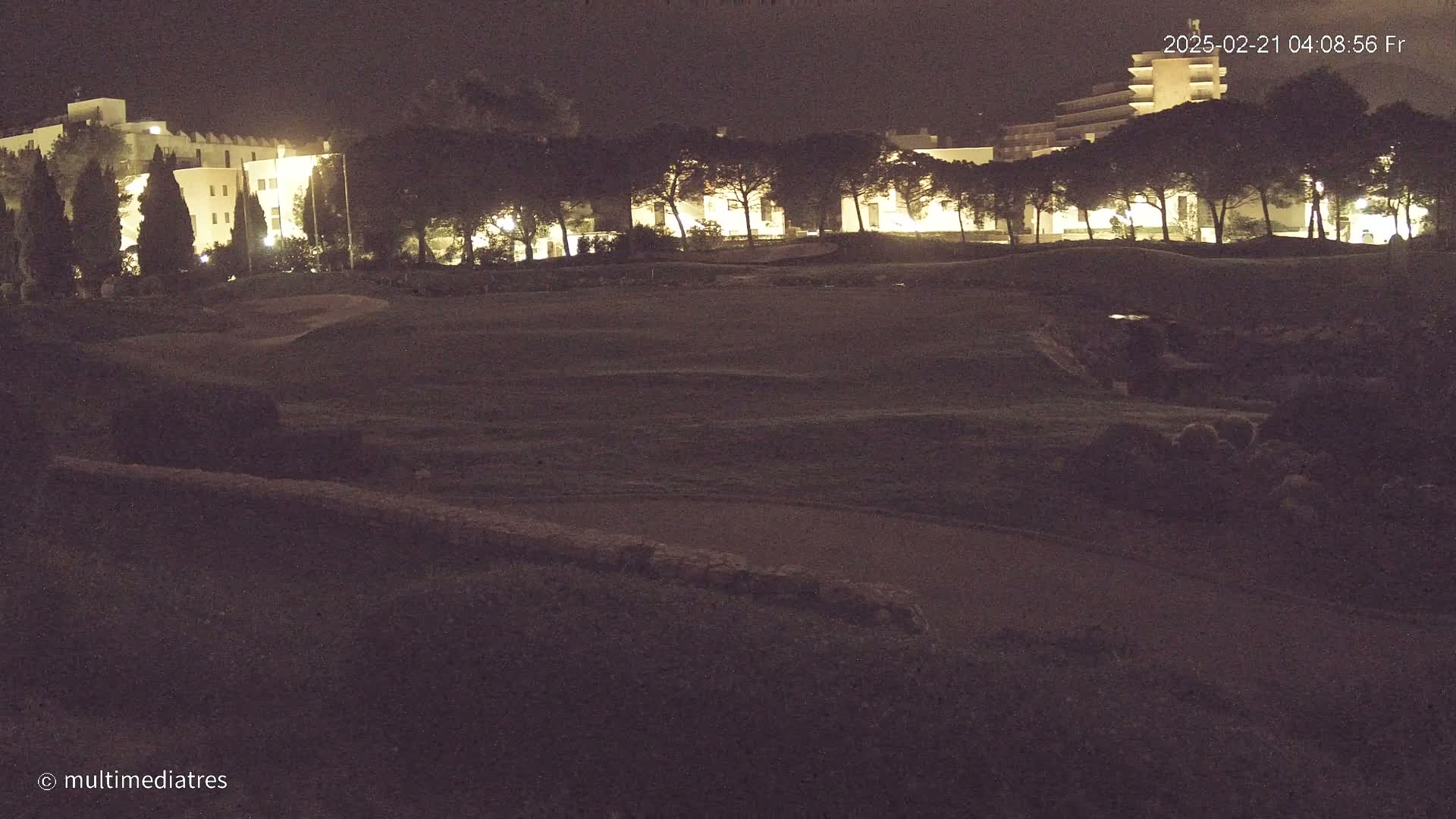 A dark, grassy area at night is illuminated by the lights of a building and trees behind it.