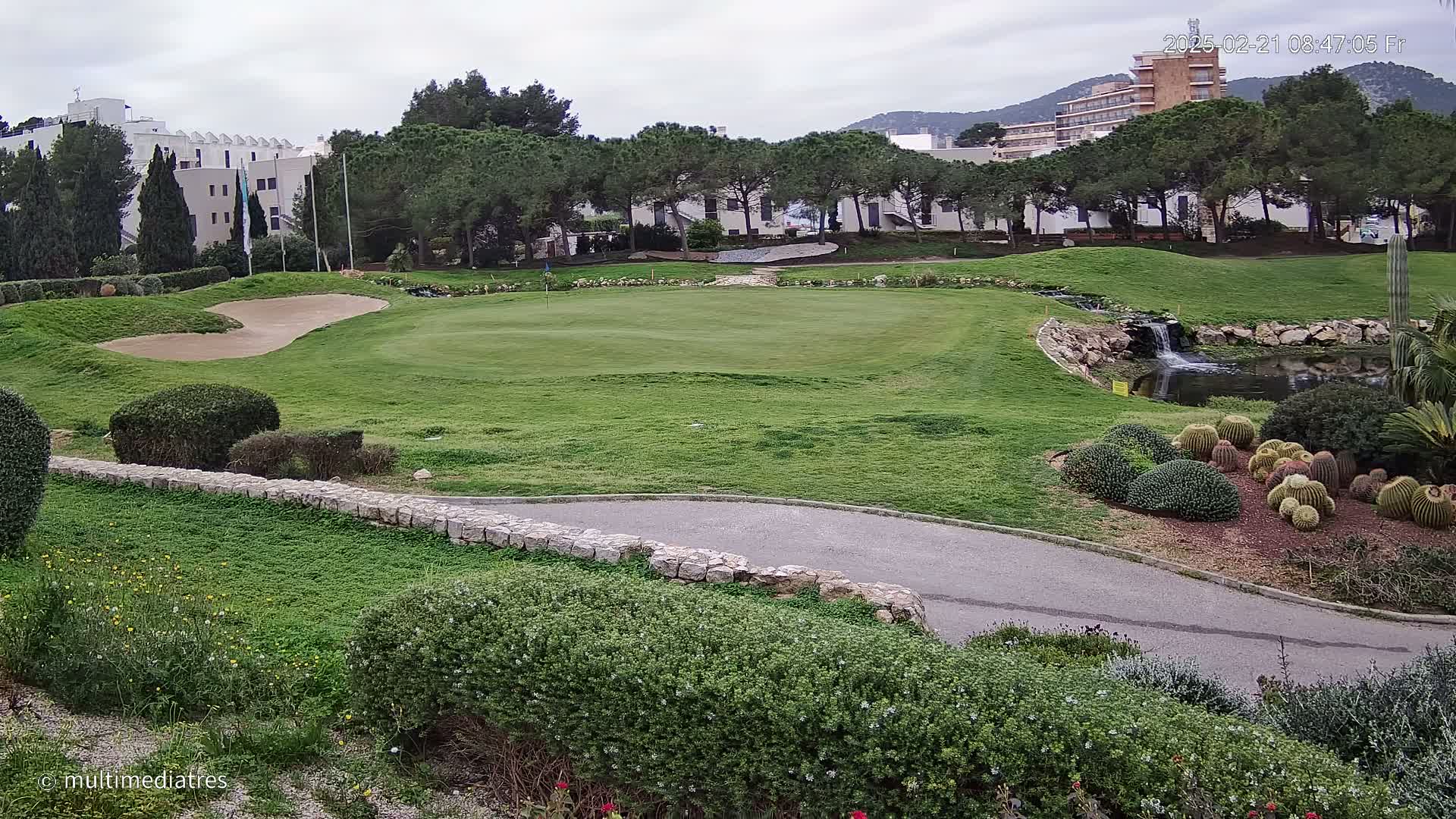 A golf course green with a sand trap, a small waterfall and pond, and a collection of cacti is seen on an overcast day, with buildings in the background.