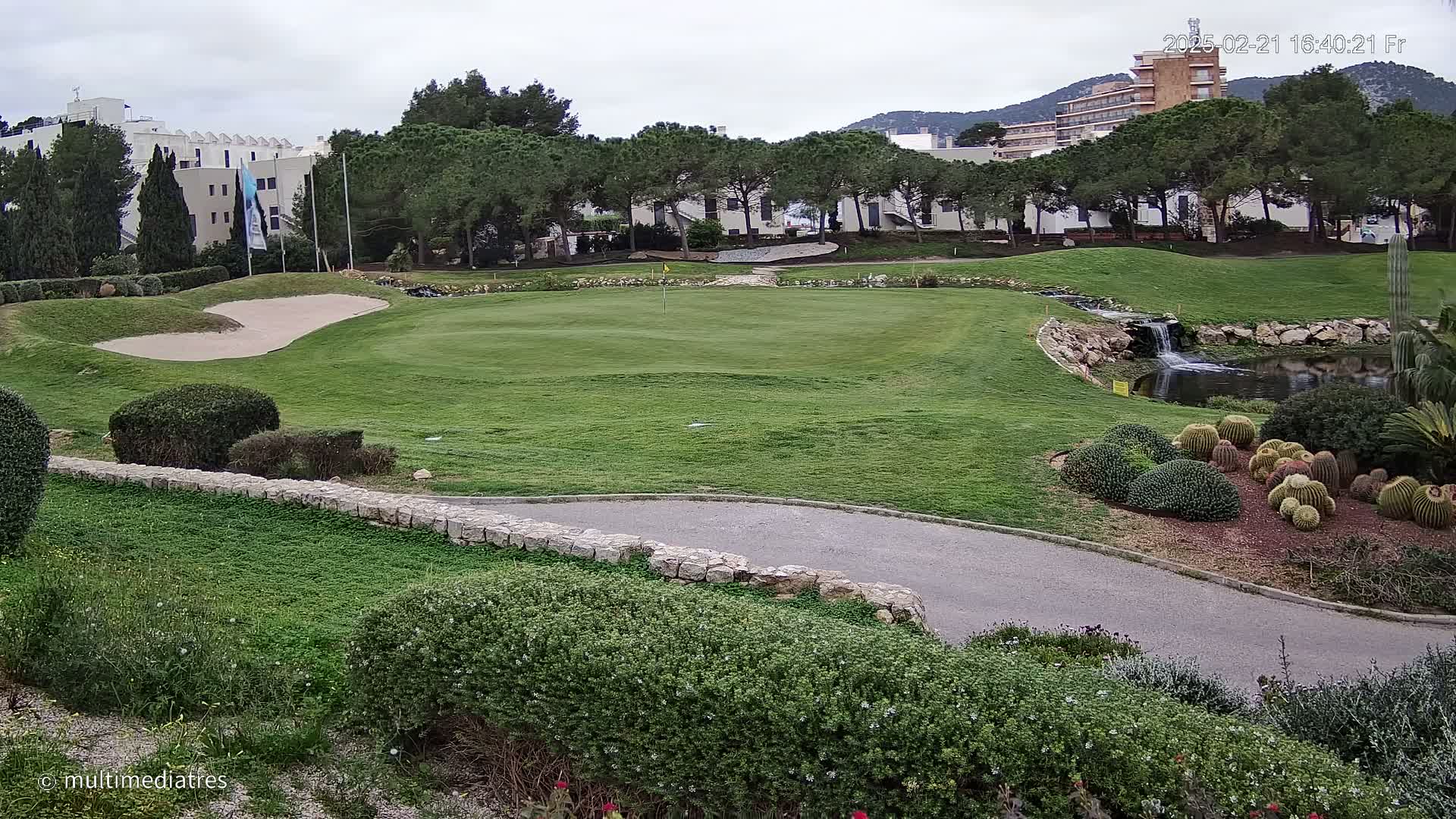 A golf course green with a sand trap, a small waterfall and pond, and a cluster of cacti, all under an overcast sky.