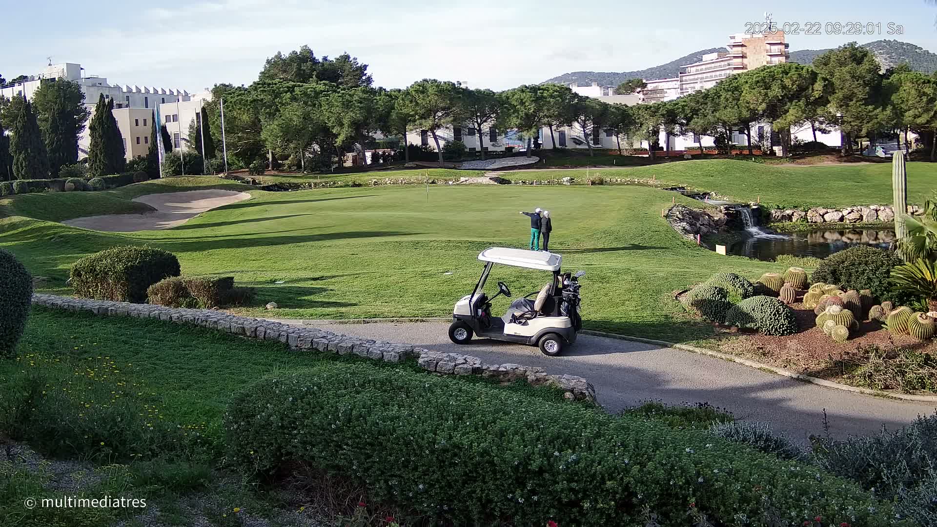A golf course with a golf cart and two people standing on a green, under a partly sunny sky.