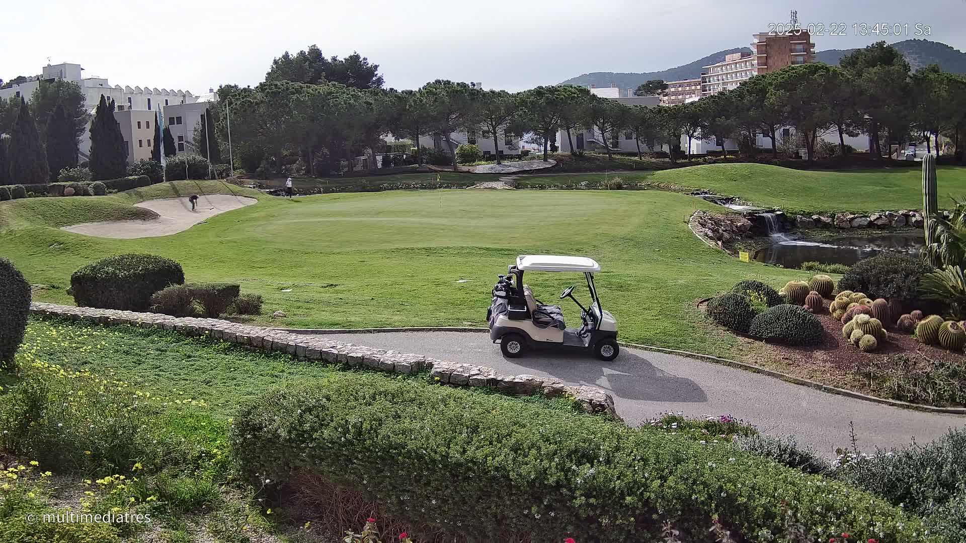 A golf course with a golf cart parked on a path, a sand trap, a small waterfall, and buildings in the background under a mostly cloudy sky.