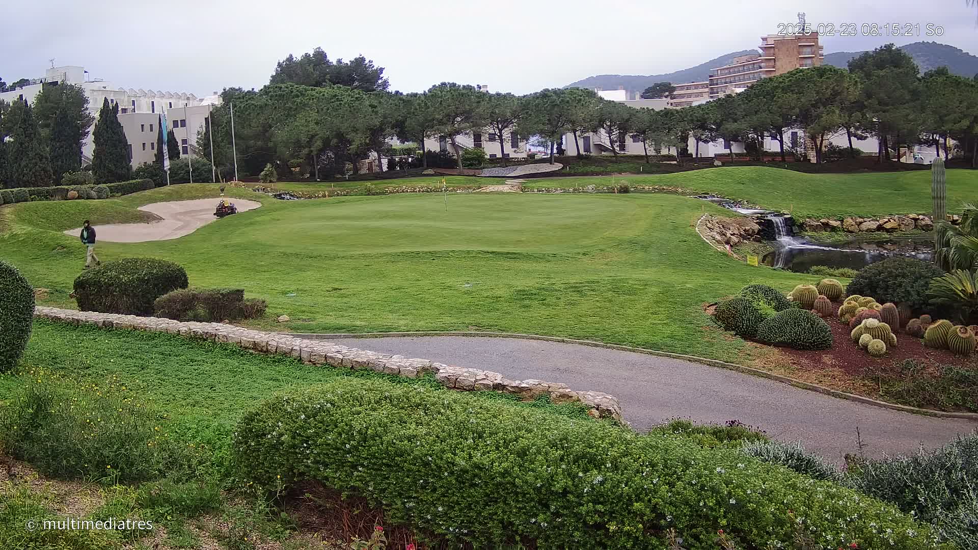 A golf course with a green, sand trap, small waterfall, and a person walking on a cloudy day.