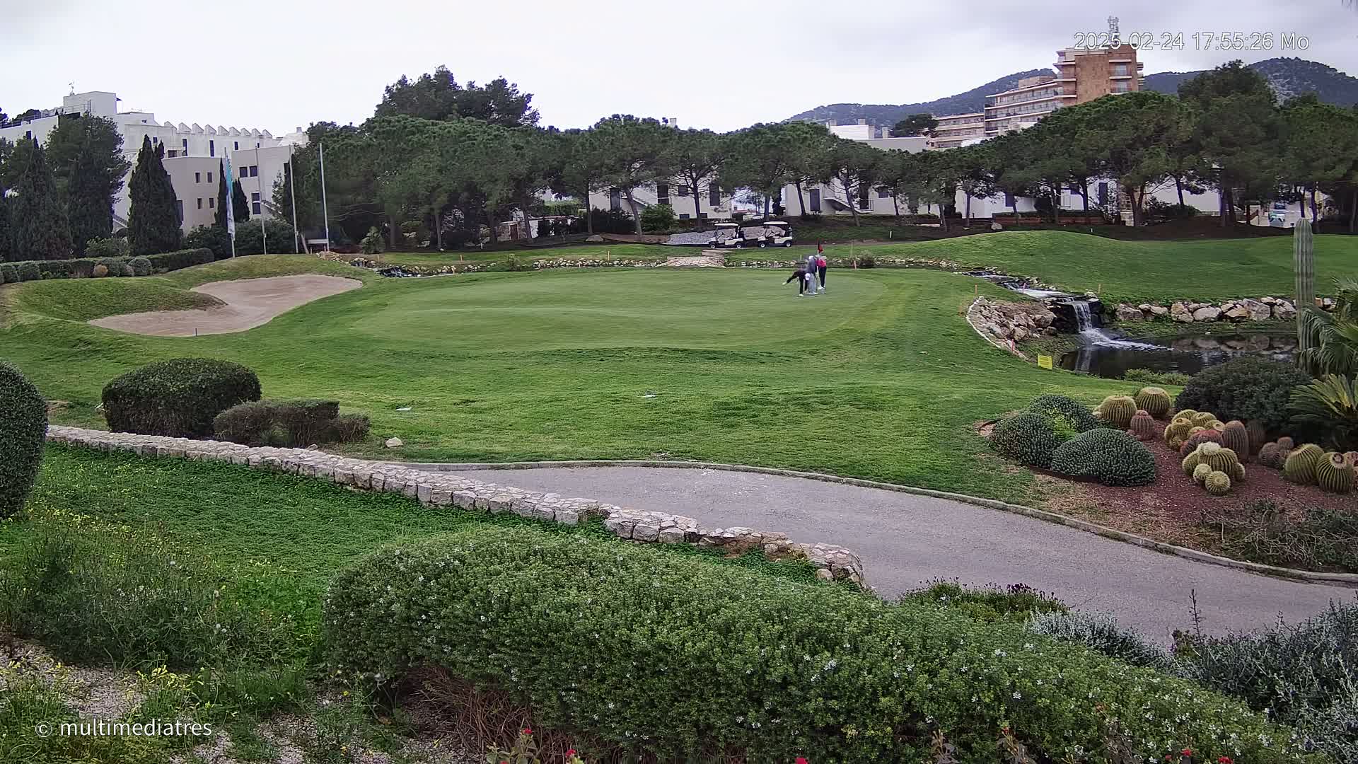 A golf course with a putting green, sand trap, small waterfall, and a group of golfers under an overcast sky.