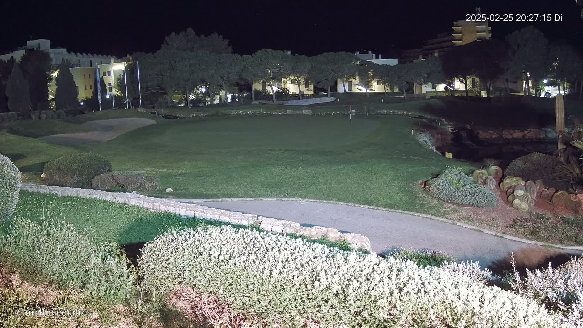 A lit golf course at night, bordered by low shrubs and trees, with a building visible in the background.