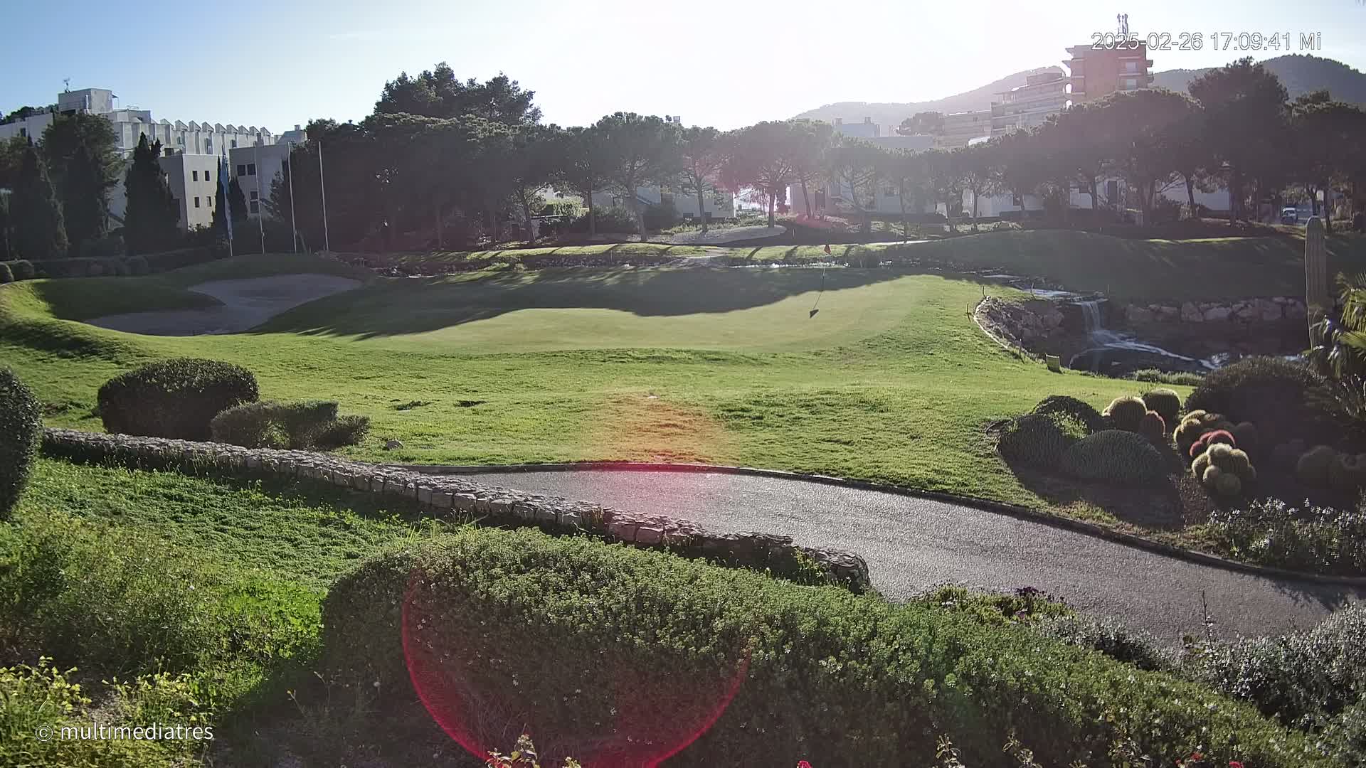 A sunny view of a golf course green with a paved path, landscaped areas, and buildings in the background.