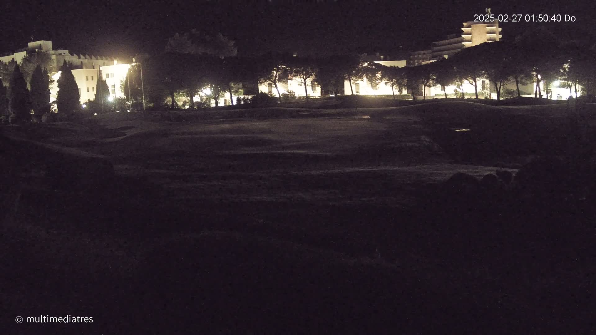 At night, illuminated buildings stand behind a dark, grassy area, possibly a golf course, under a clear sky.