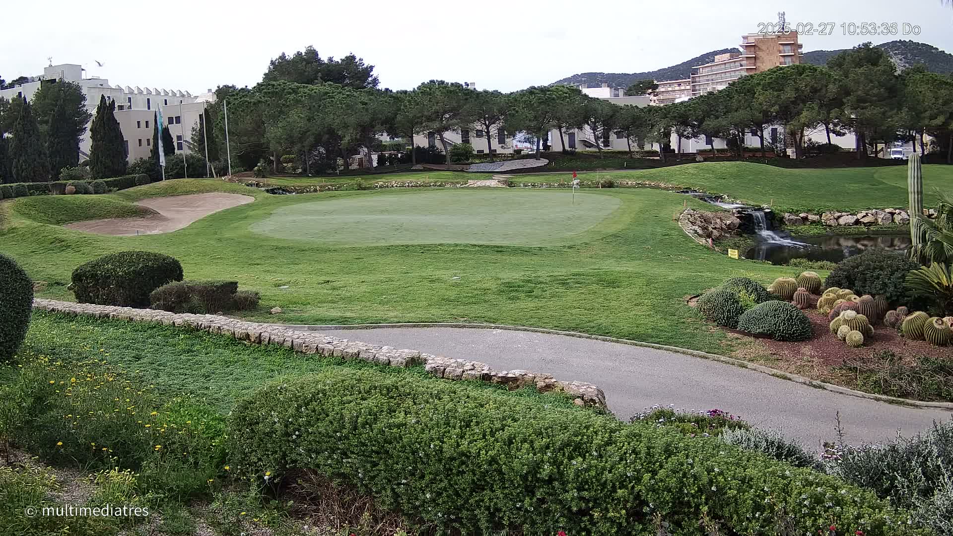 A golf course putting green with a small pond and waterfall is situated near a residential building complex under an overcast sky.