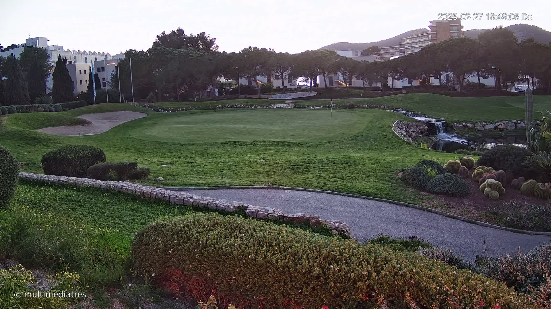 A golf course green with a sand trap, a small waterfall, and a paved path is visible under a twilight sky, near buildings and lush greenery.