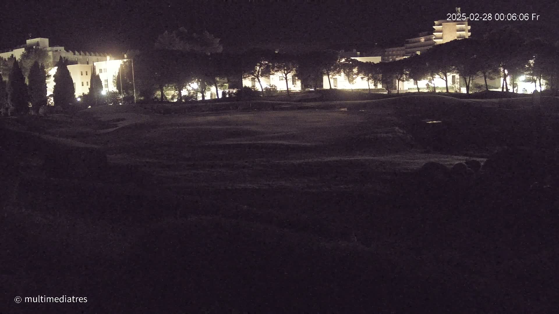 A nighttime view of a dark, grassy area with illuminated buildings and trees in the background under a clear, dark sky.