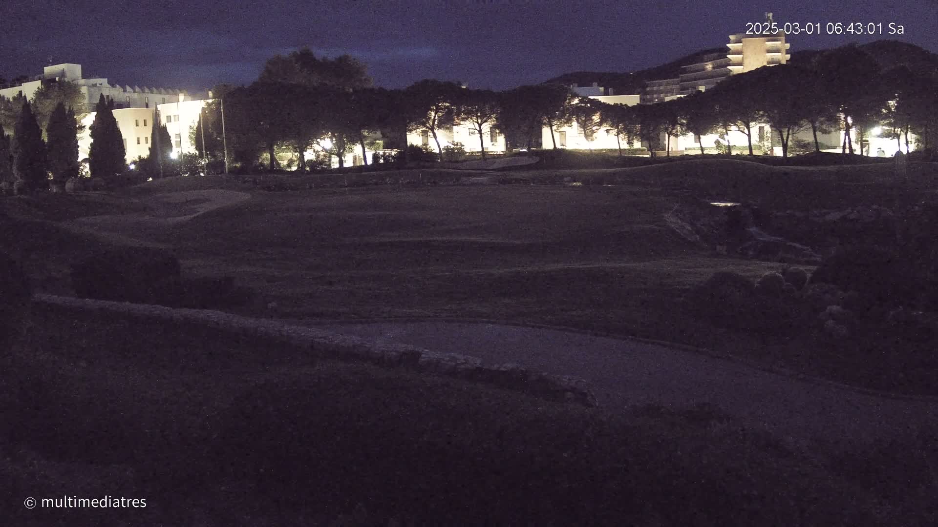 At night, a dark, grassy area with a building complex in the background is illuminated by artificial light.