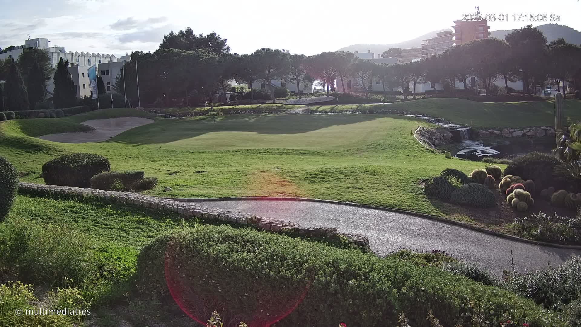 A sunny view of a golf course green with a sand trap, a small waterfall, and landscaping featuring shrubs and cacti.
