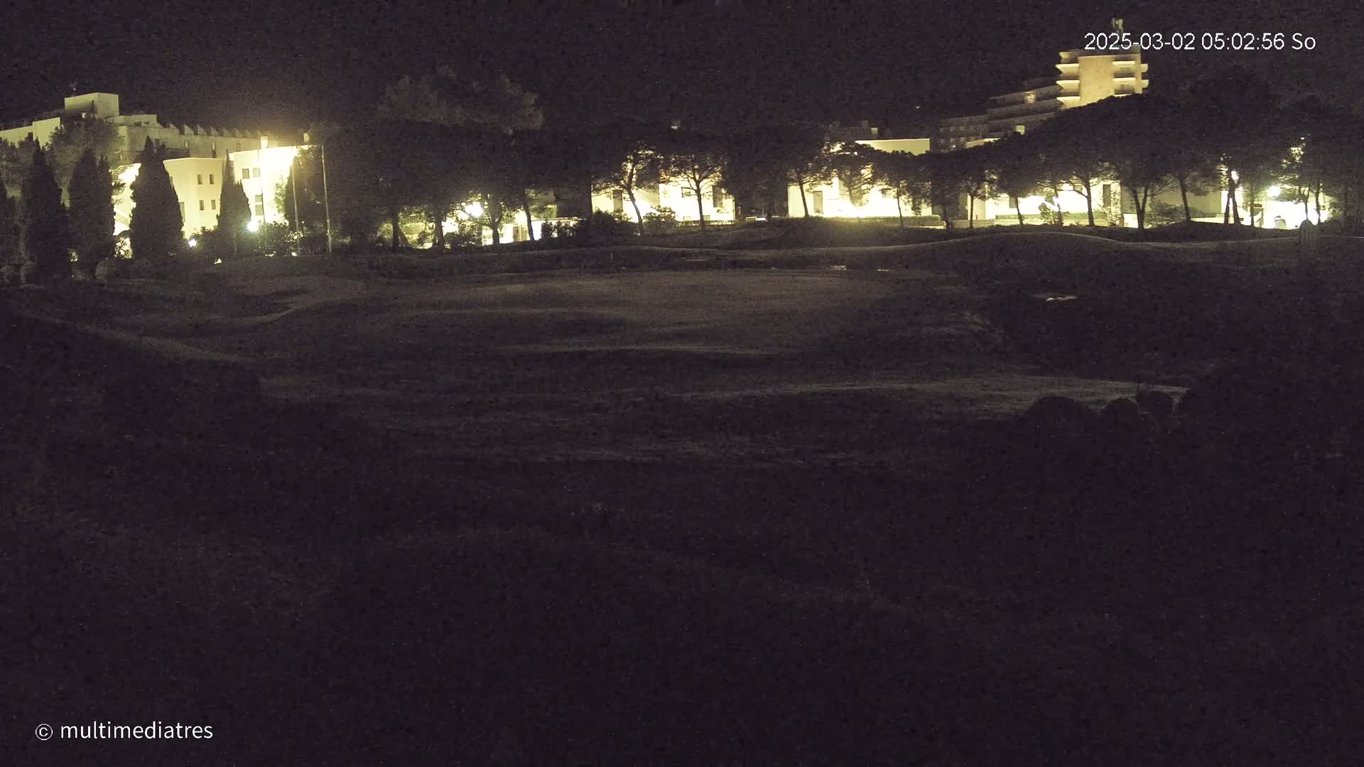 At night, illuminated buildings are visible behind a dark, grassy area.