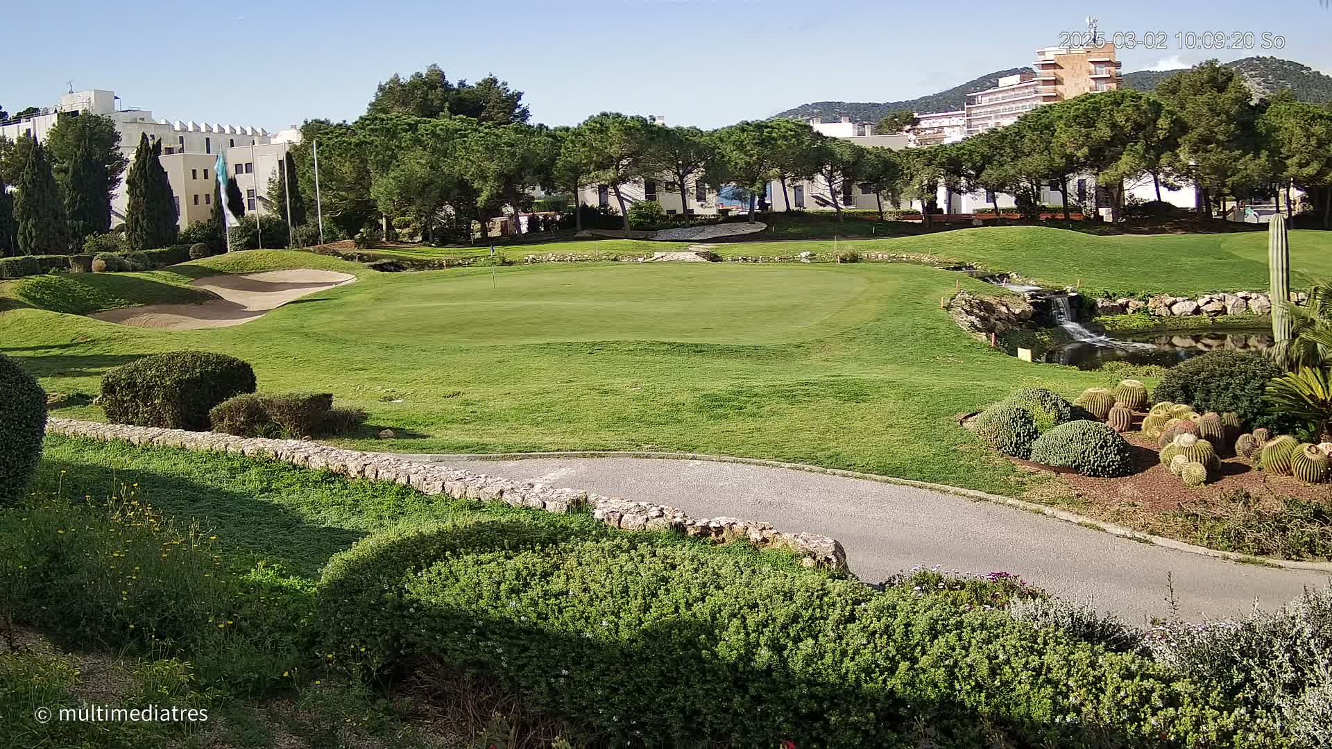A sunny golf course with a small pond and a waterfall, surrounded by manicured lawns, shrubs, and cacti, sits in front of buildings and a mountain in the distance.