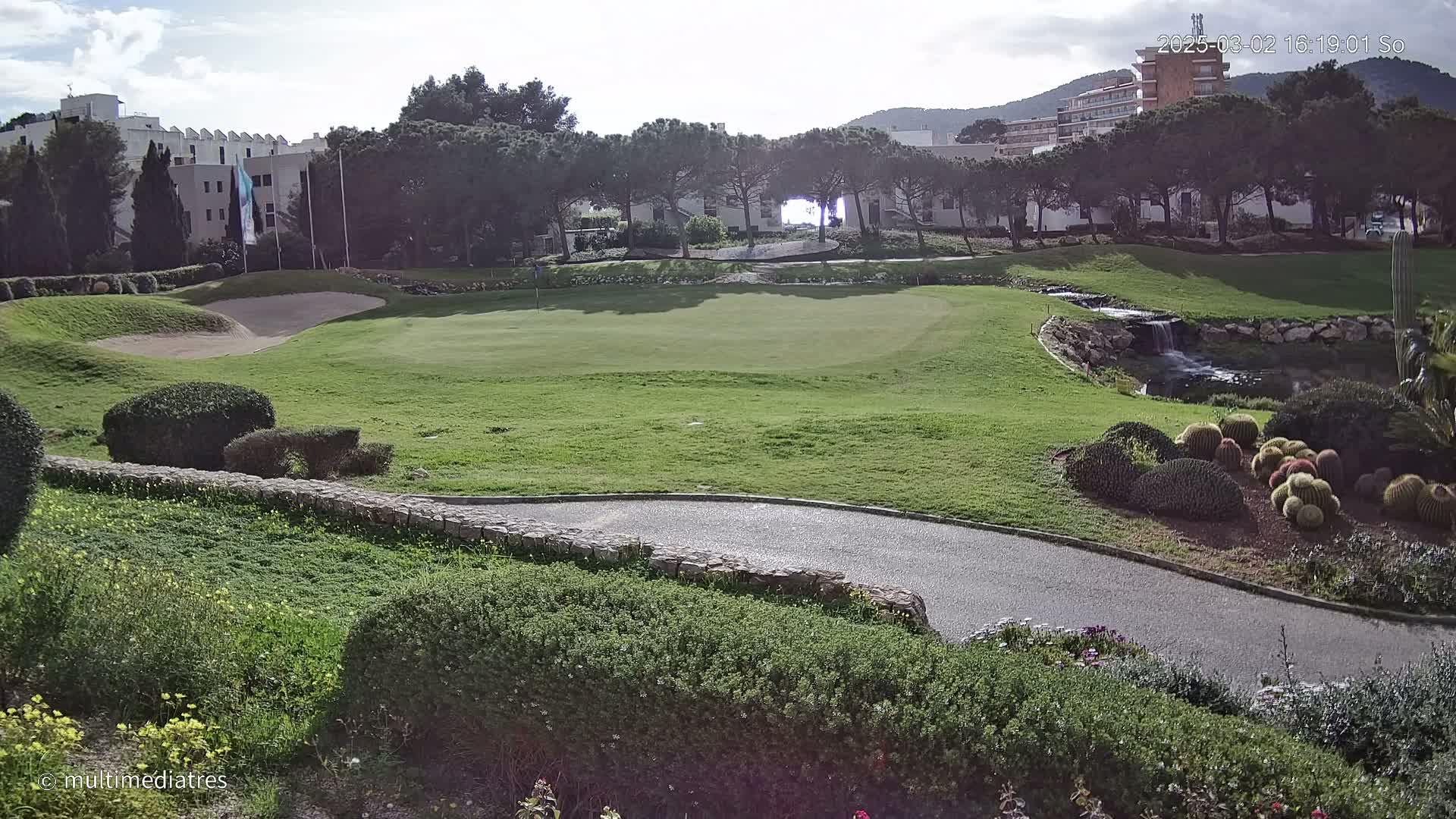 A sunny golf course with a sand trap, a small waterfall, and a variety of shrubs and cacti, situated near buildings and trees.