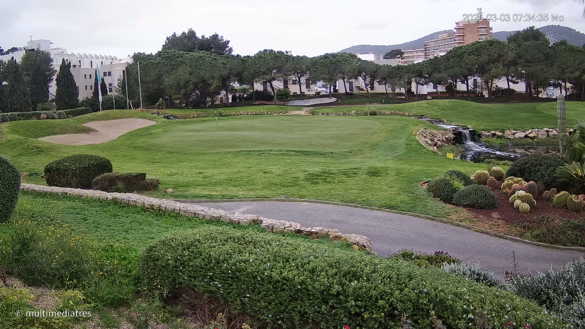 A golf course green with a sand trap, a small waterfall, and a cluster of cacti under an overcast sky.