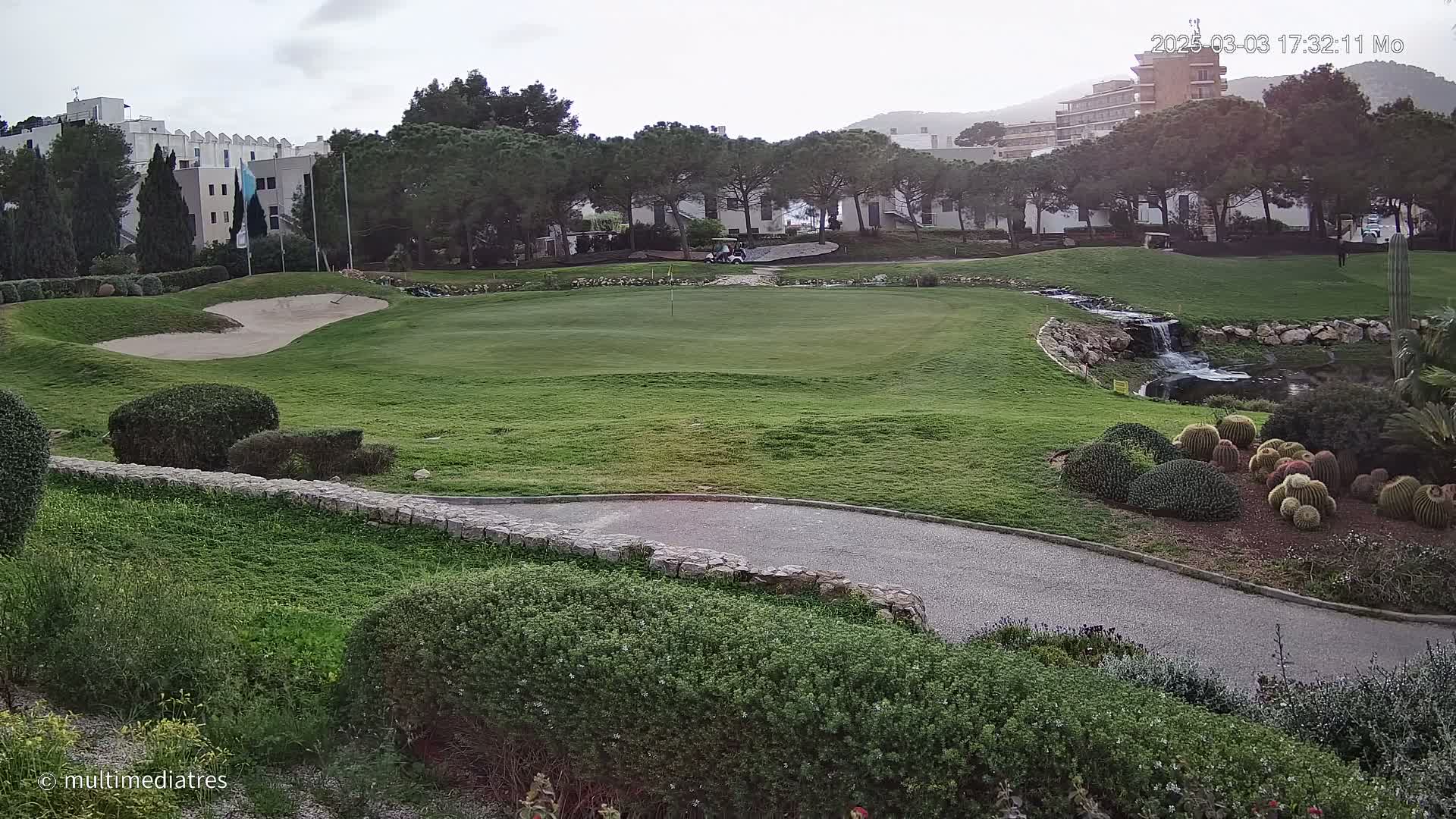 A golf course green with a sand trap, a small waterfall, and a golf cart is visible under an overcast sky.