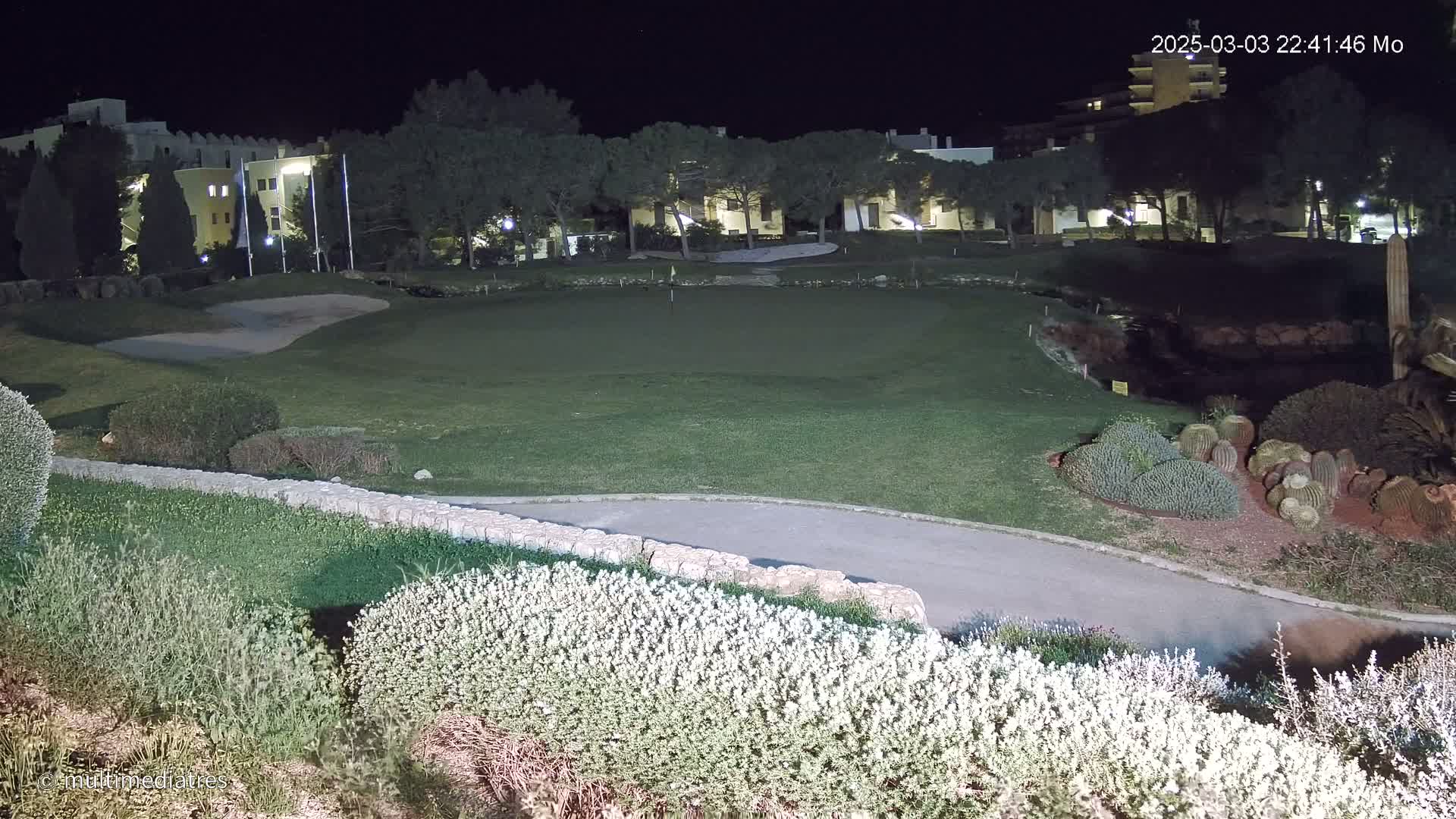 A nighttime view of a golf course, illuminated by artificial lights, with buildings and trees visible in the background.