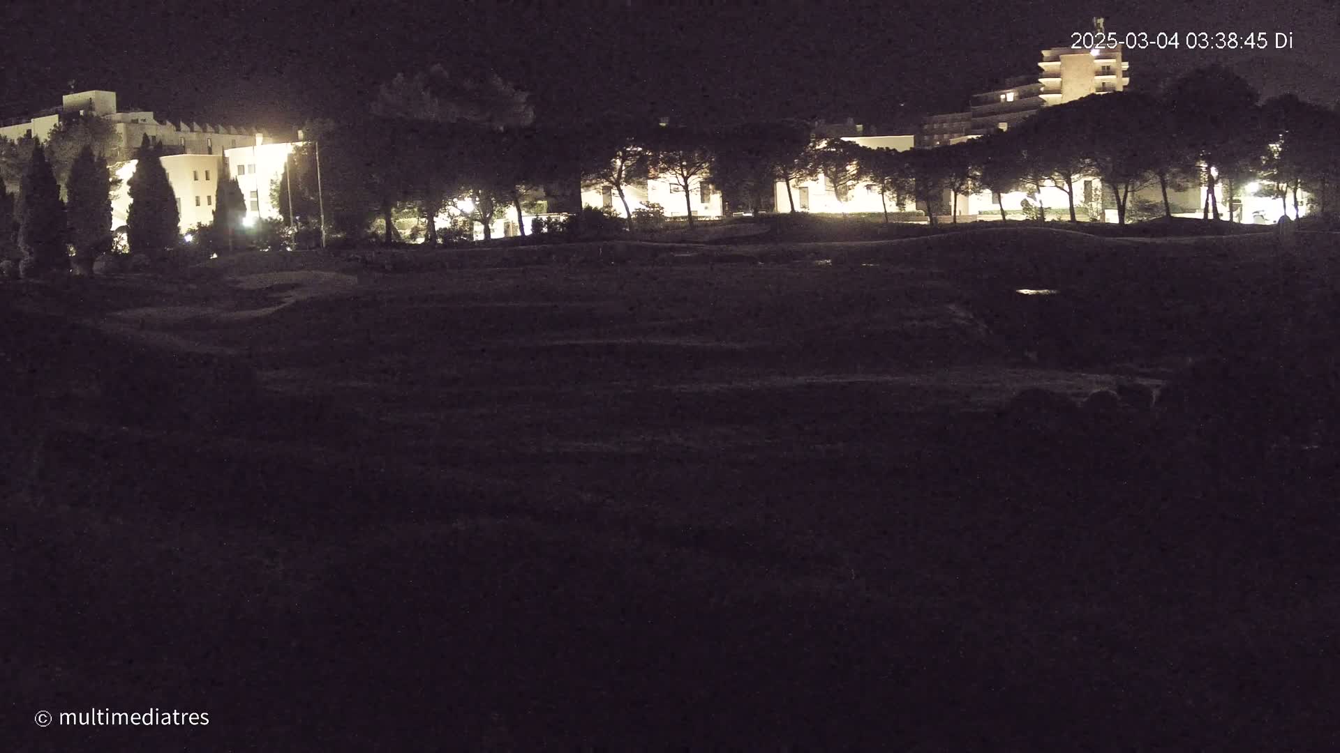 At night, illuminated buildings are visible behind a dark, empty field with some trees.