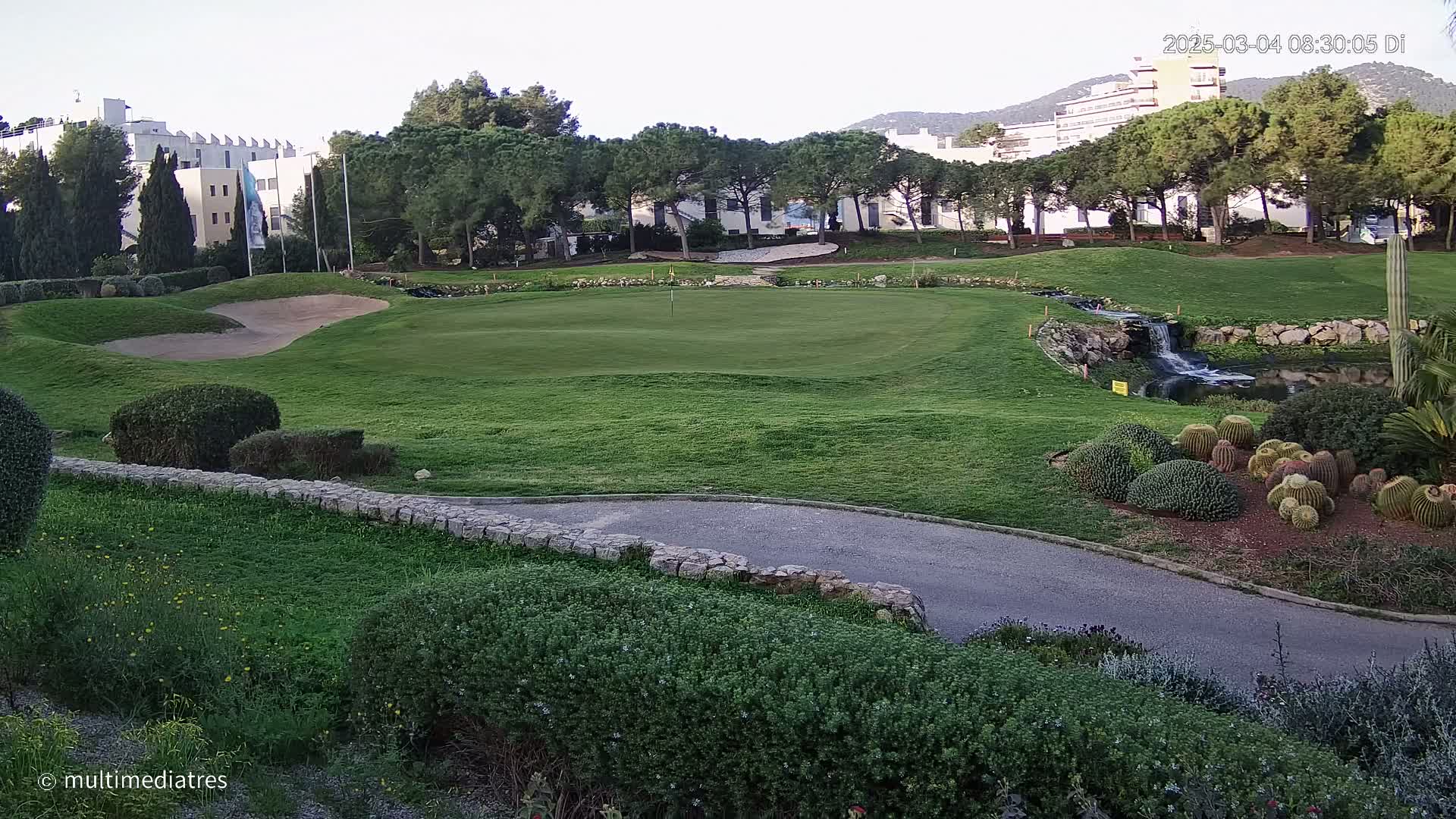 A golf course green with a sand trap, a small waterfall, and a collection of cacti is visible under a clear sky.