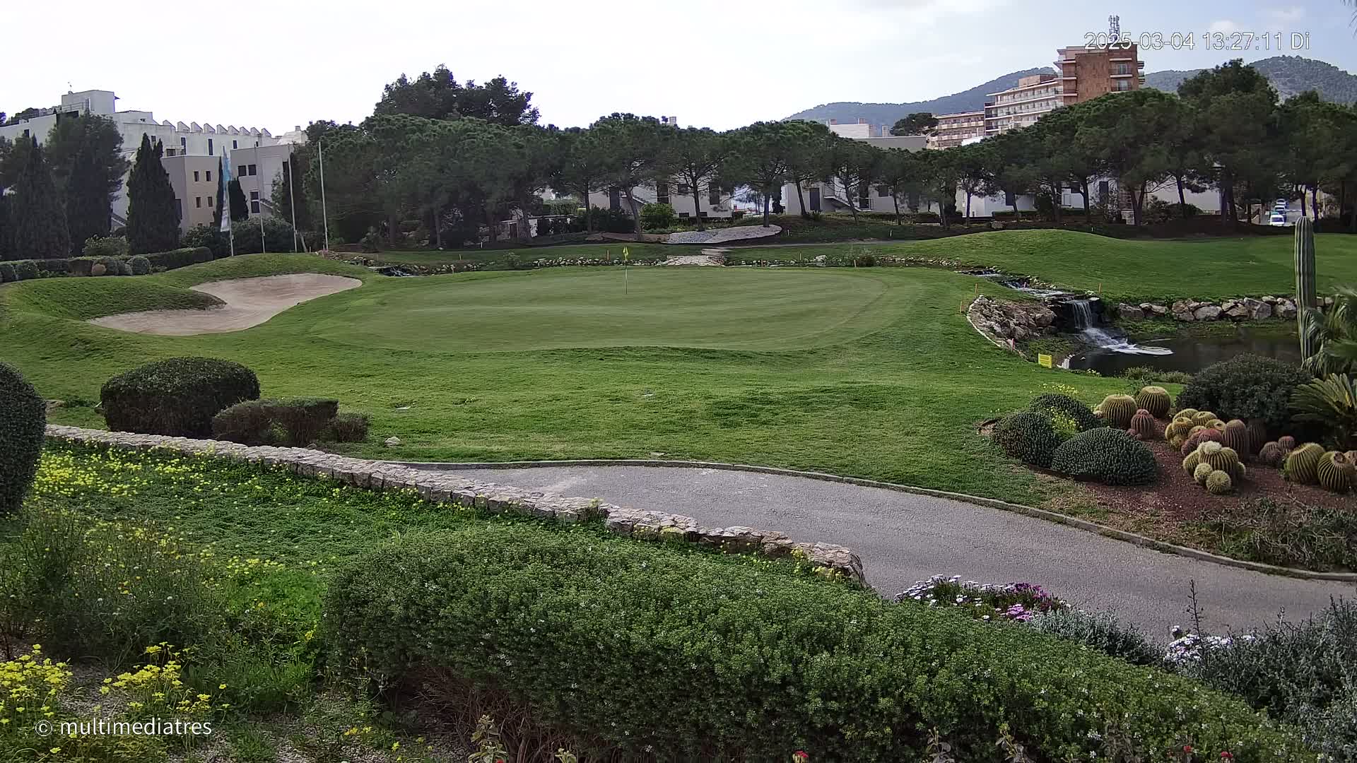 A golf course green with a sand trap, a small waterfall, and a variety of cacti and shrubs, all under an overcast sky.