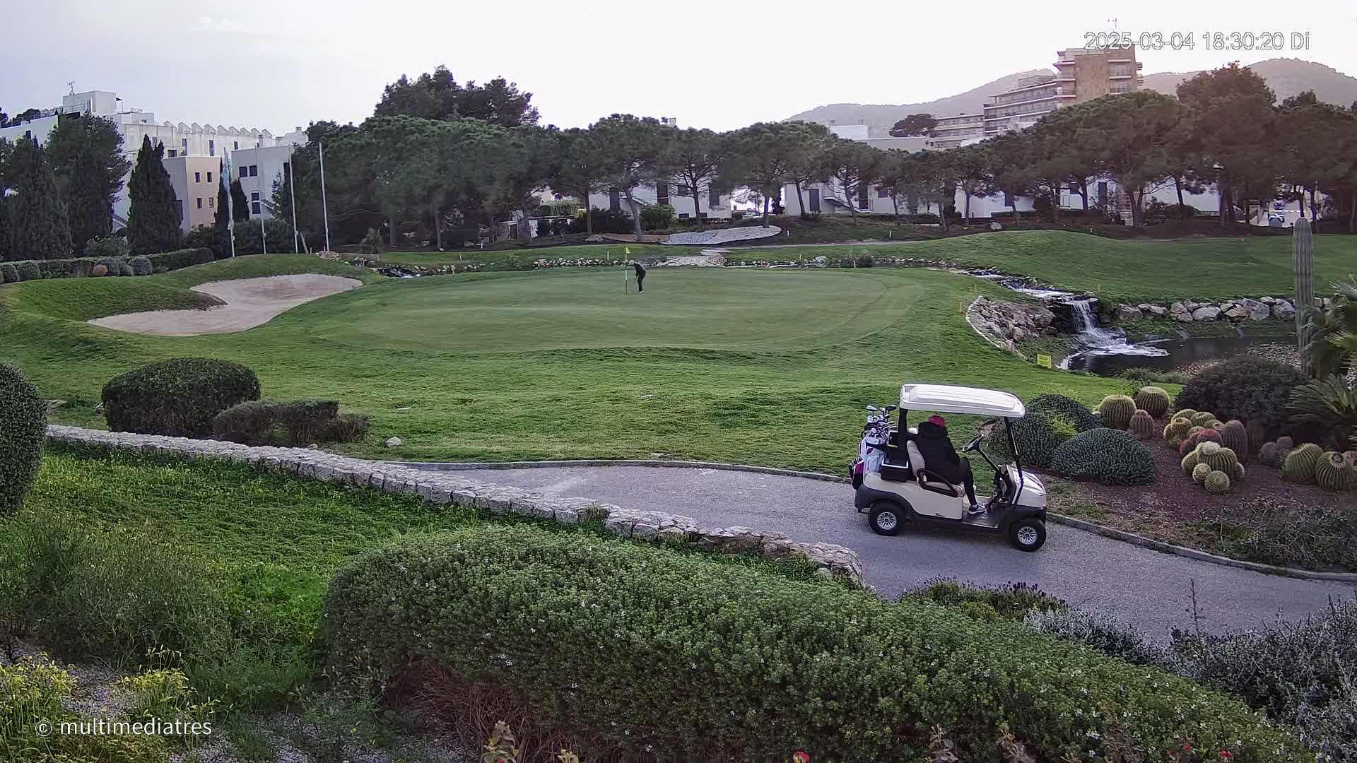 A golf course with a golfer putting on the green, a golf cart on a path, and a small waterfall in the background under an overcast sky.