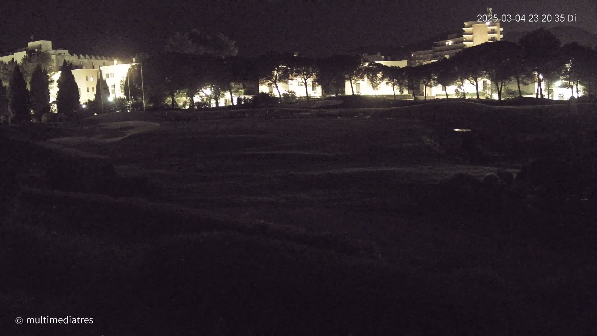 At night, a row of illuminated buildings is visible behind a dark expanse of land, possibly a field or golf course, lined with dark trees.