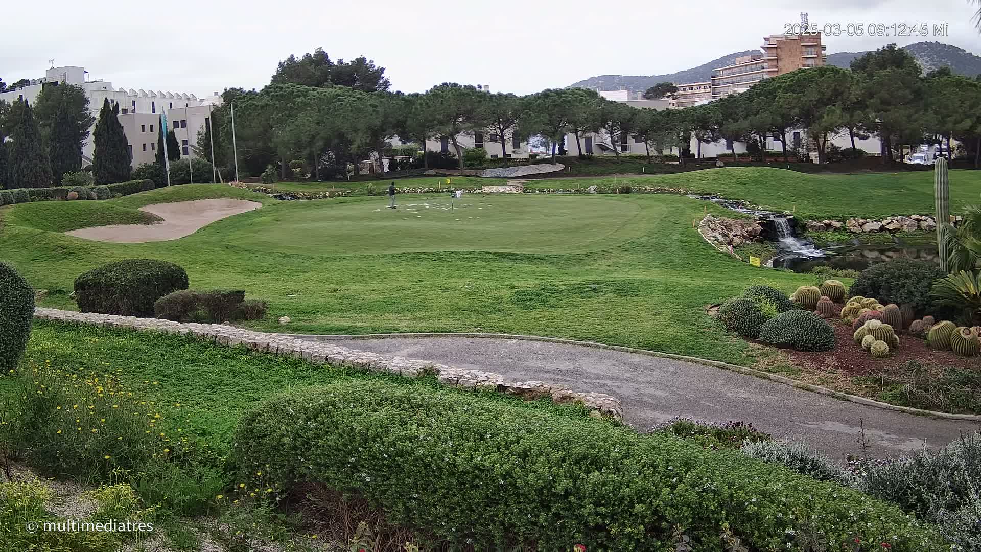 A golf course putting green with a sand trap, a small waterfall, and a collection of cacti is visible, under an overcast sky.