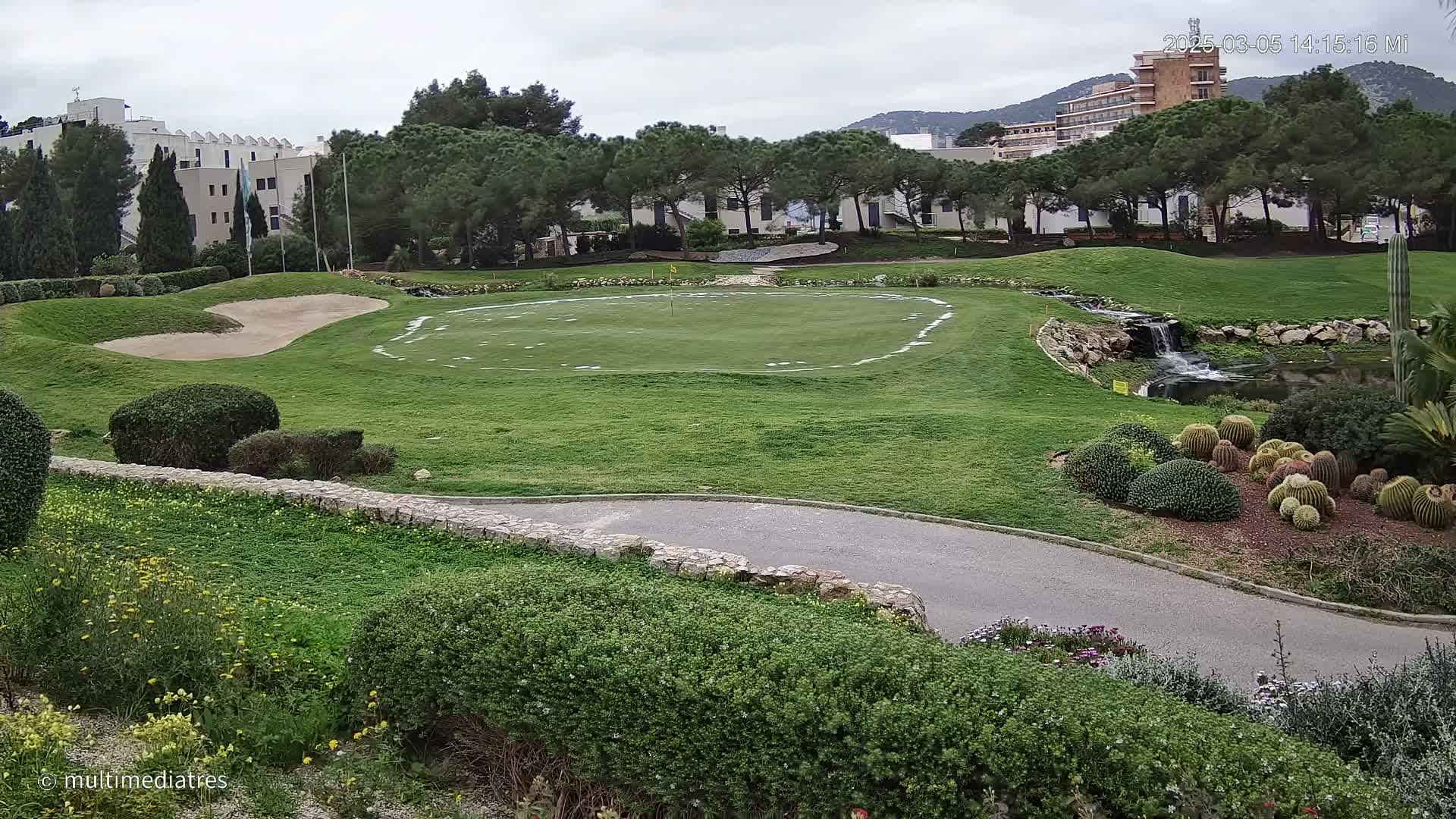 A putting green on a golf course is surrounded by landscaping, including a small waterfall and cacti, with buildings and trees in the background under an overcast sky.