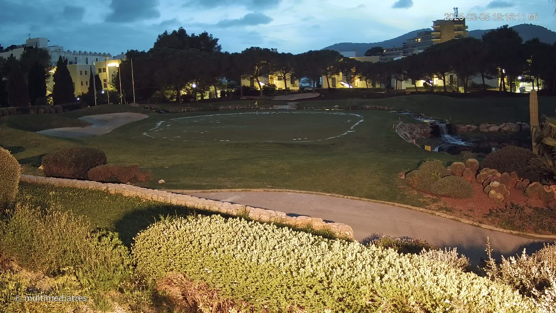A putting green at dusk is surrounded by landscaping, a small waterfall, and buildings in the background under a twilight sky.