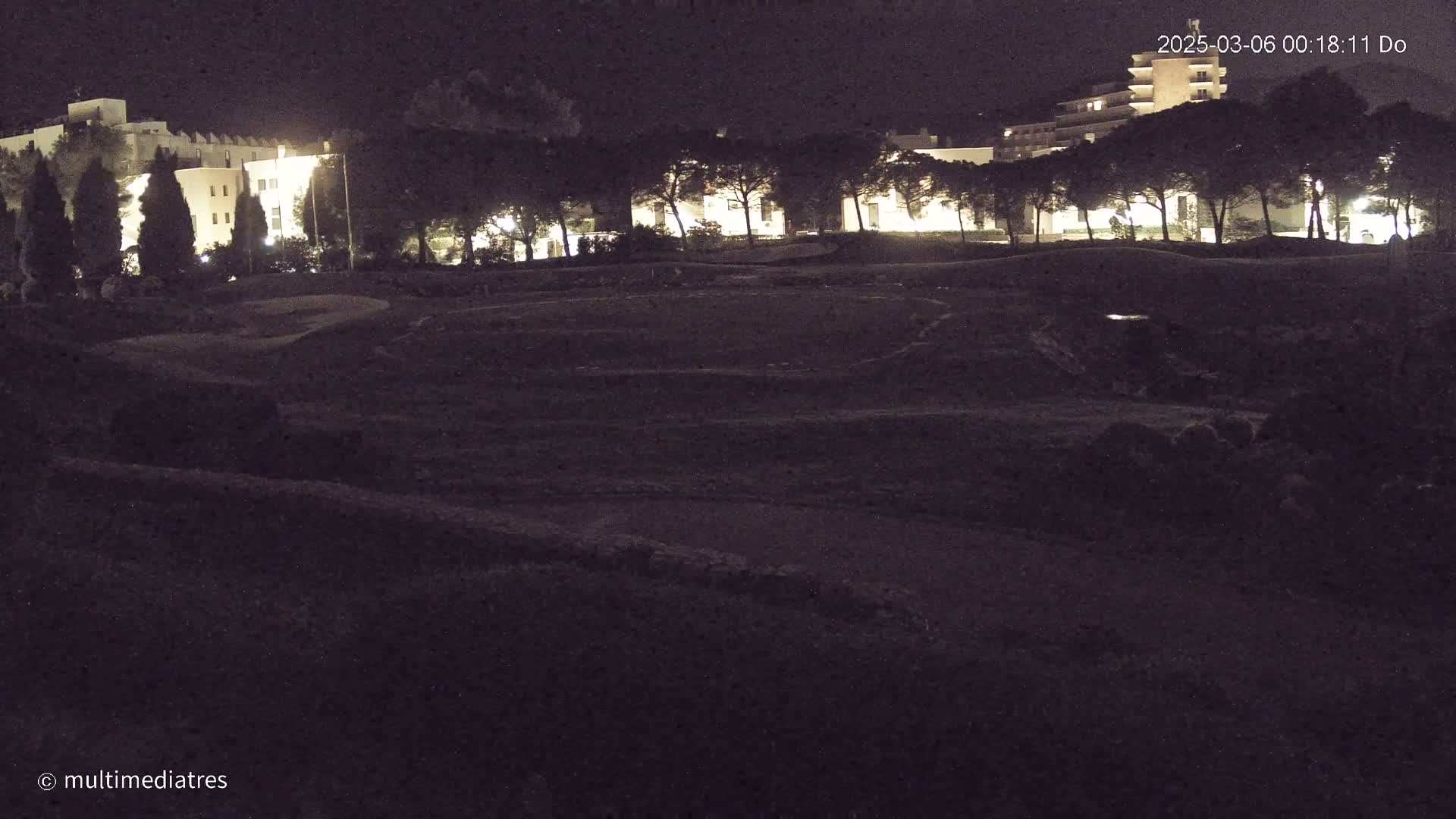 A dark, nighttime view of a grassy area with a row of illuminated buildings and trees in the background.