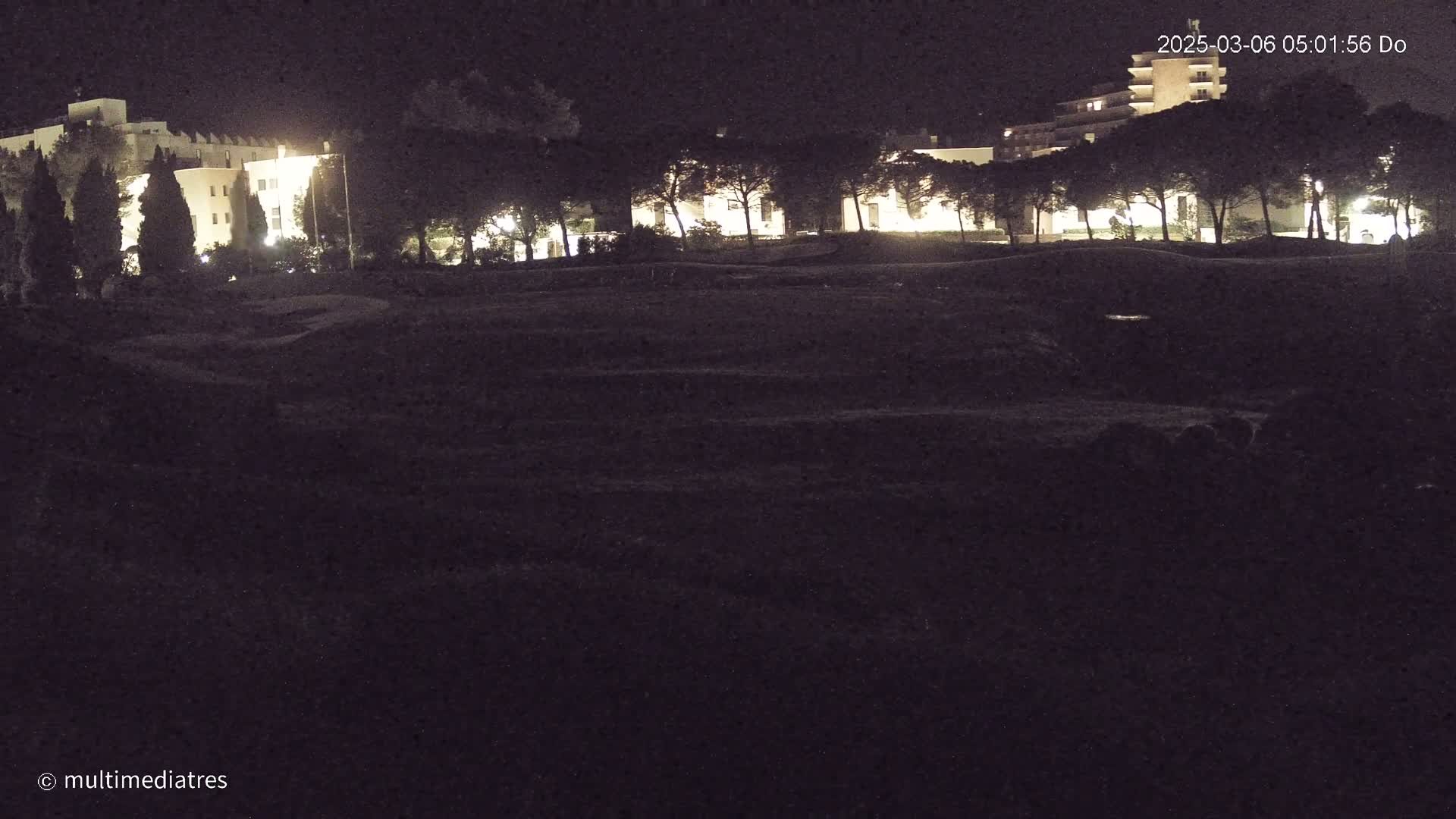 At night, illuminated buildings are visible behind a line of trees, overlooking a dark, mostly flat expanse of land.