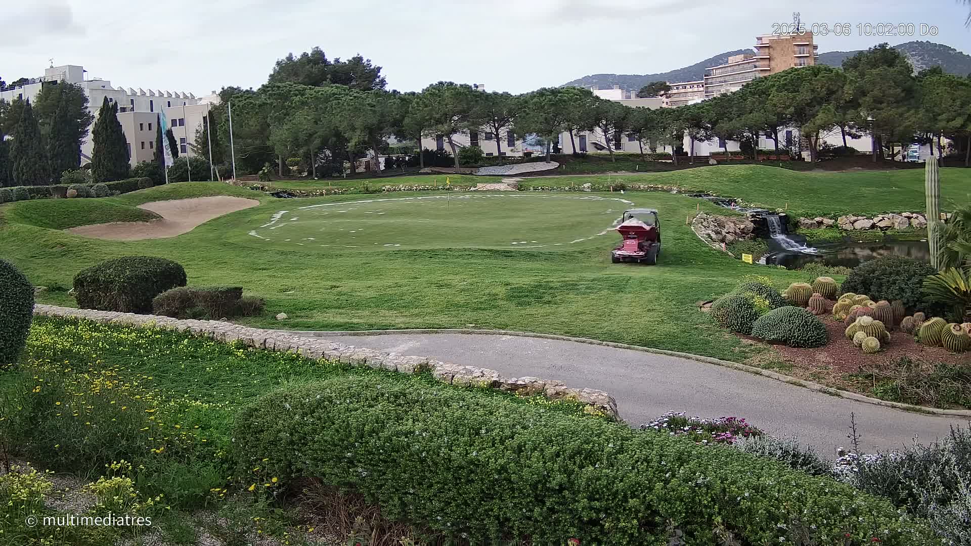 A putting green on a golf course, with a small vehicle, is surrounded by landscaping, a small waterfall, and buildings in the background under an overcast sky.