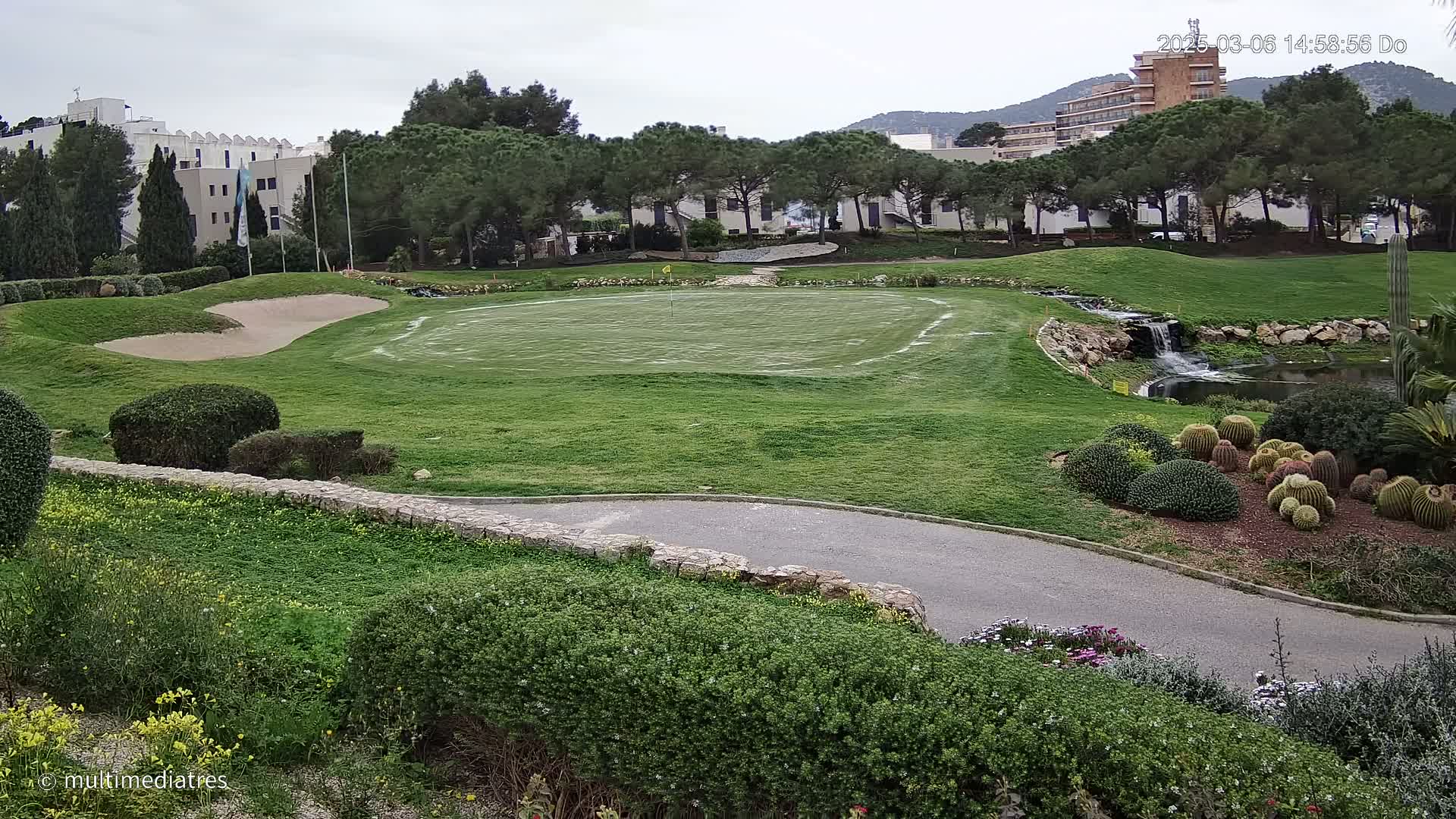 A golf course putting green with a sand trap and a small waterfall under an overcast sky.