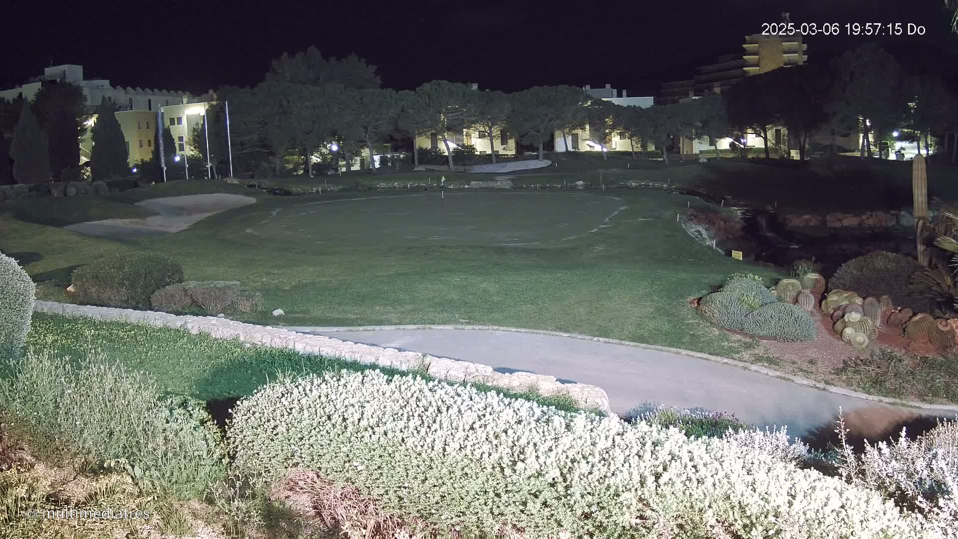 A nighttime view of a putting green illuminated by artificial light, surrounded by landscaping, with buildings and trees in the background.