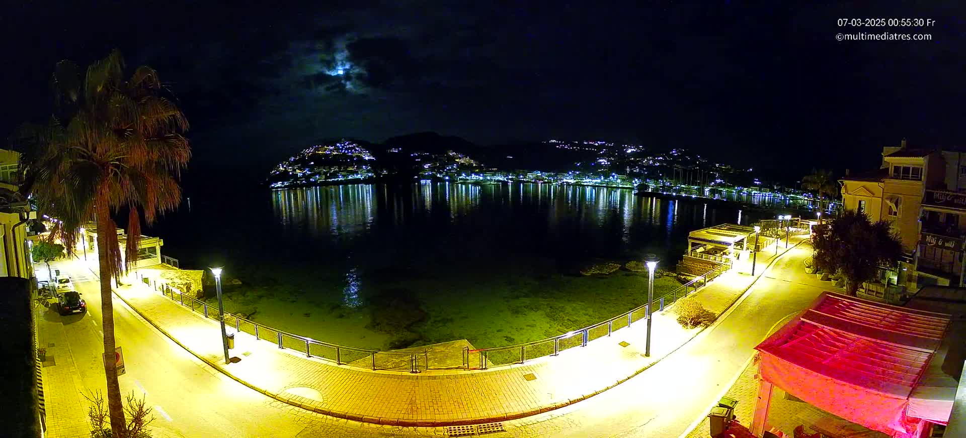 A moonlit night scene shows a calm body of water reflecting the lights of a town nestled on a hillside, with a paved waterfront path illuminated by lampposts.