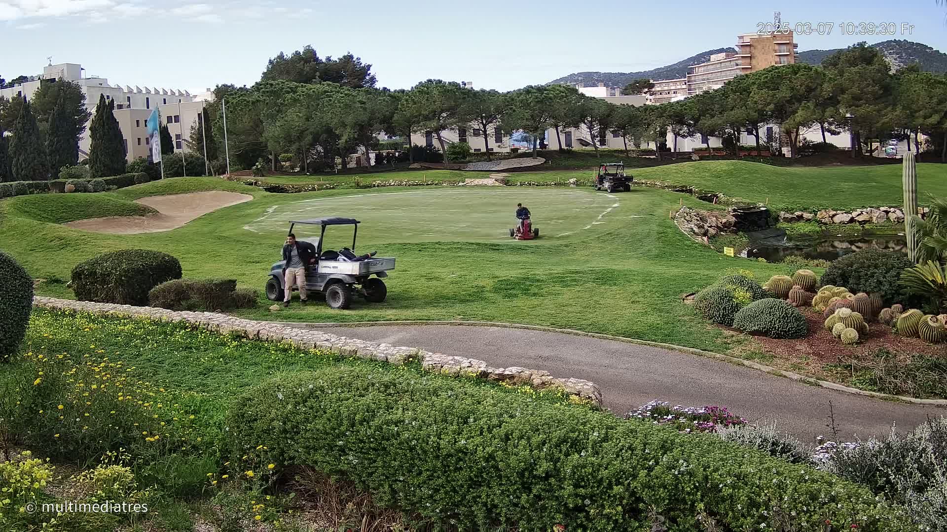 A golf course with several workers and golf carts, under a mostly clear sky.