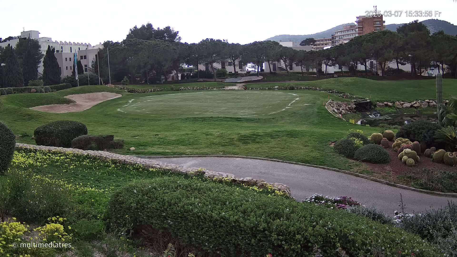 A golf course putting green with sand traps, landscaping, and buildings in the background under an overcast sky.