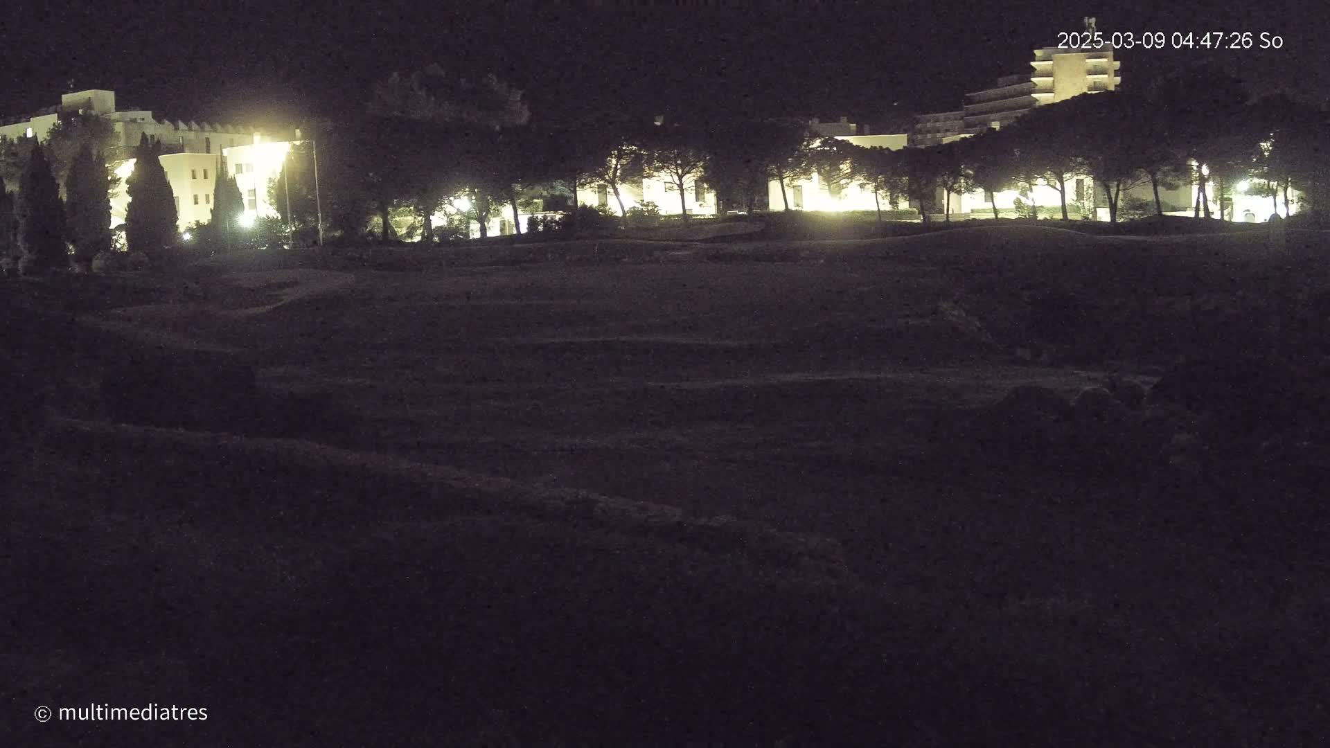 At night, illuminated buildings are visible beyond a dark, uneven field lined with trees.