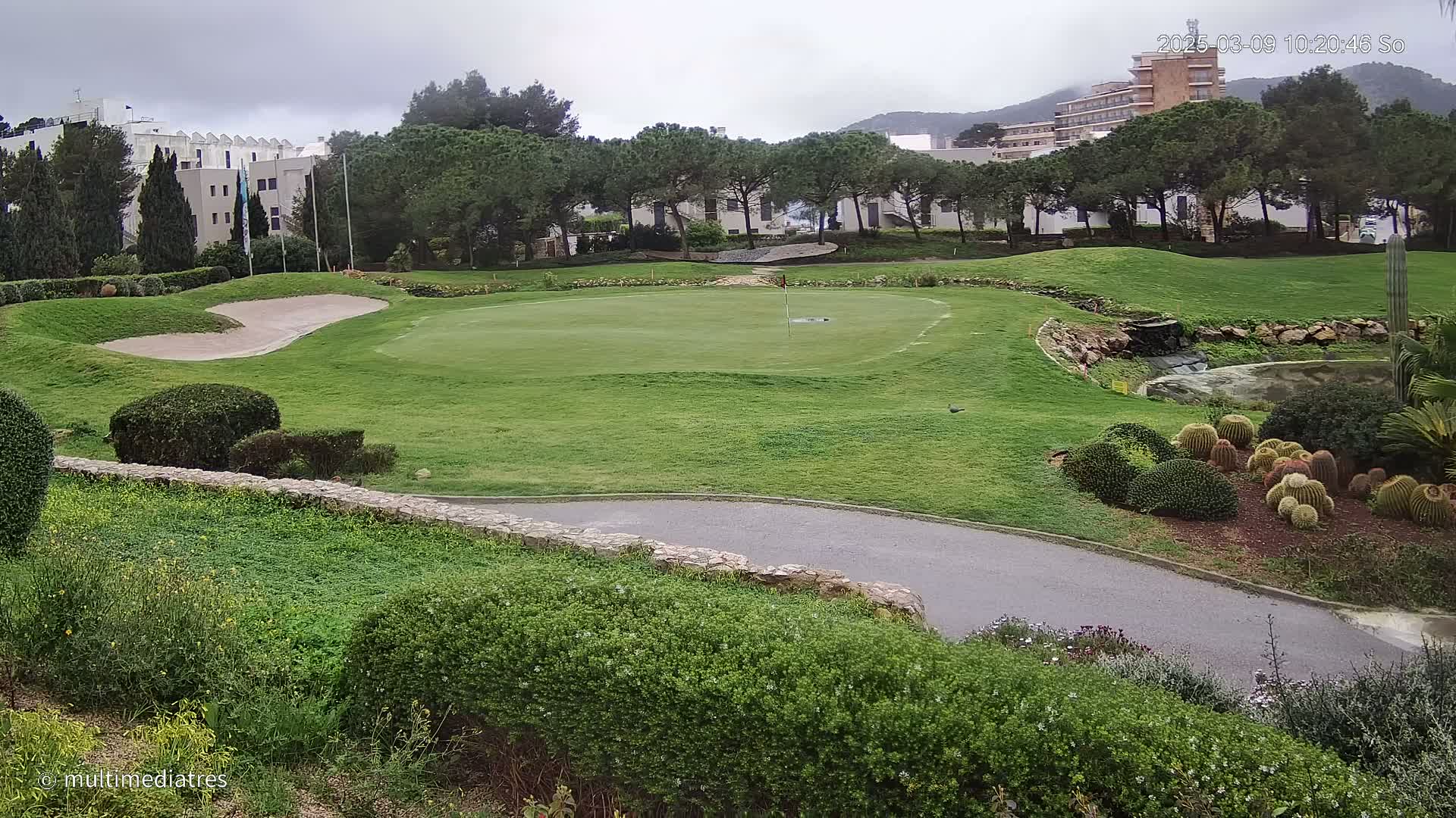 A golf course green with a sand trap and a small water feature is visible, under an overcast sky, with buildings and trees in the background.
