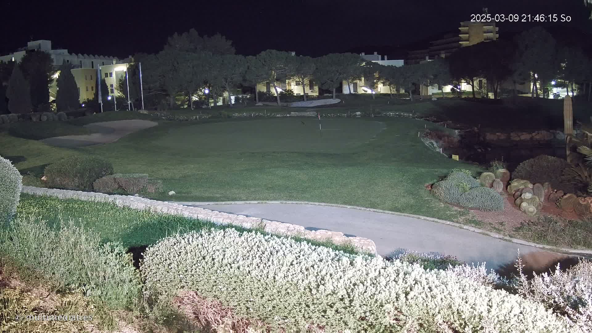 A nighttime view of a putting green on a golf course, illuminated by artificial lights, with buildings and vegetation in the background.