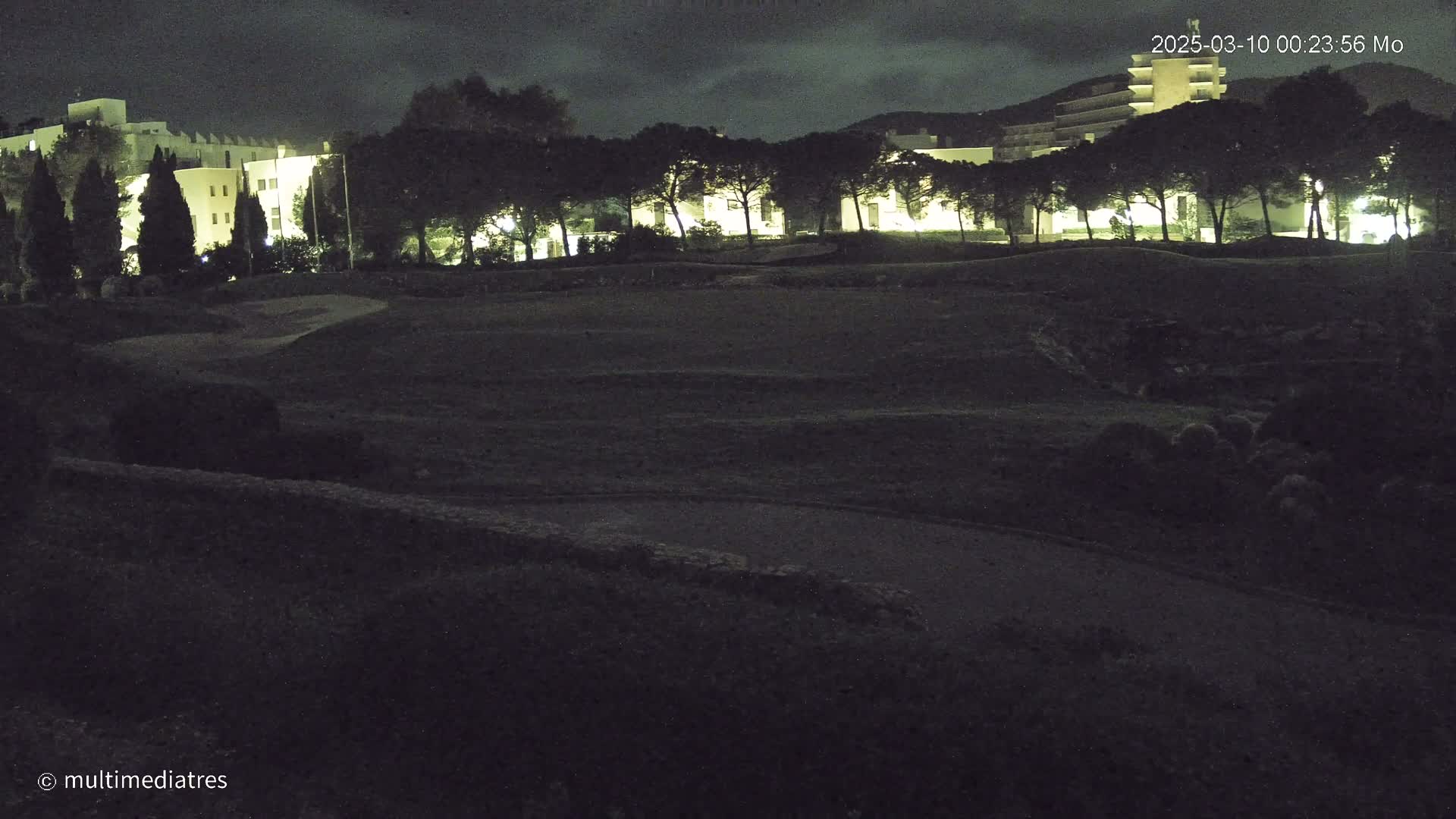 A nighttime view of a grassy area with buildings and trees in the background under a dark, cloudy sky.