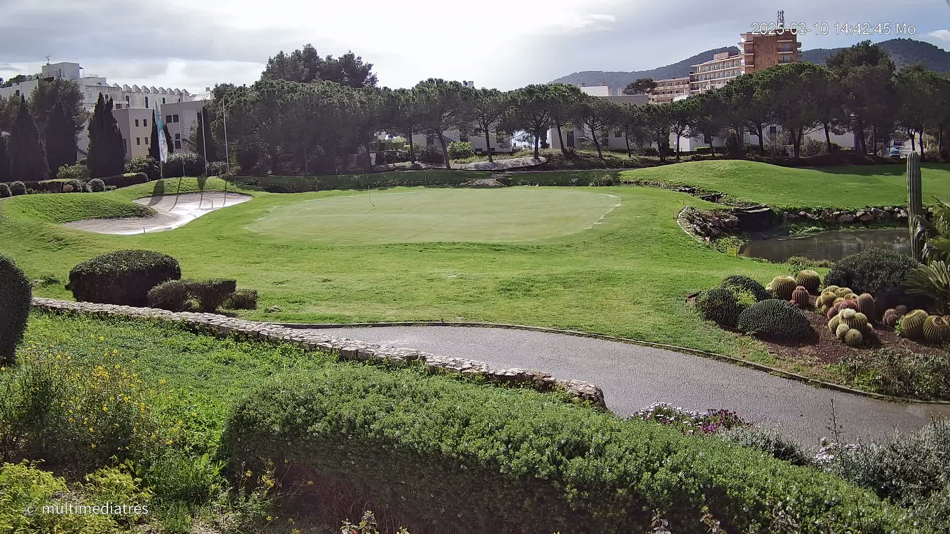 A putting green on a golf course is situated next to a low stone wall, with buildings and trees in the background under a partly cloudy sky.
