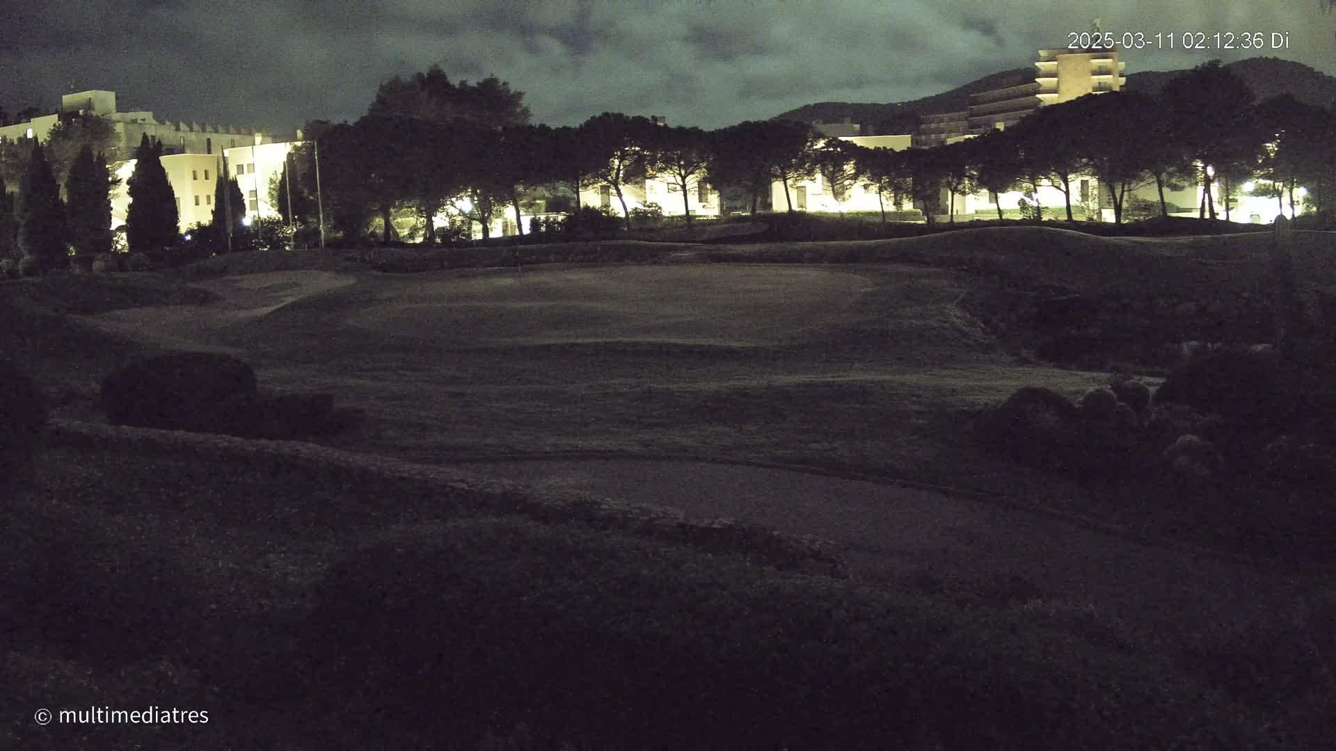 A nighttime view of a golf course with buildings and trees in the background under a cloudy sky.