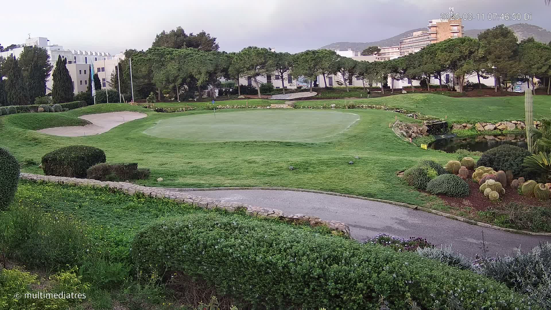 A golf course green, sand trap, and surrounding landscaping are visible under an overcast sky, with buildings and trees in the background.