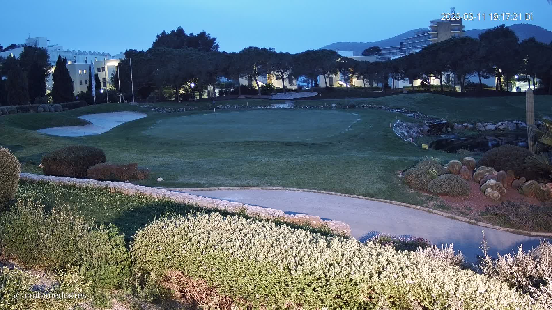 A putting green on a golf course at dusk, surrounded by landscaping, with buildings and mountains visible in the background.