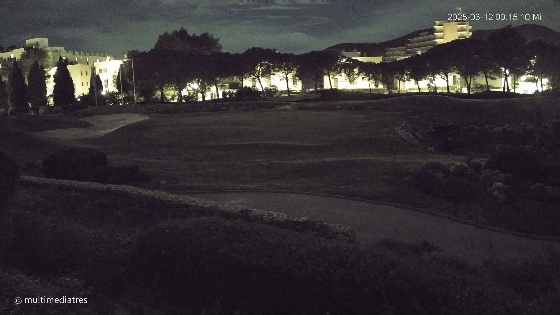 A nighttime view of a golf course with illuminated buildings and trees in the background under a partly cloudy sky.