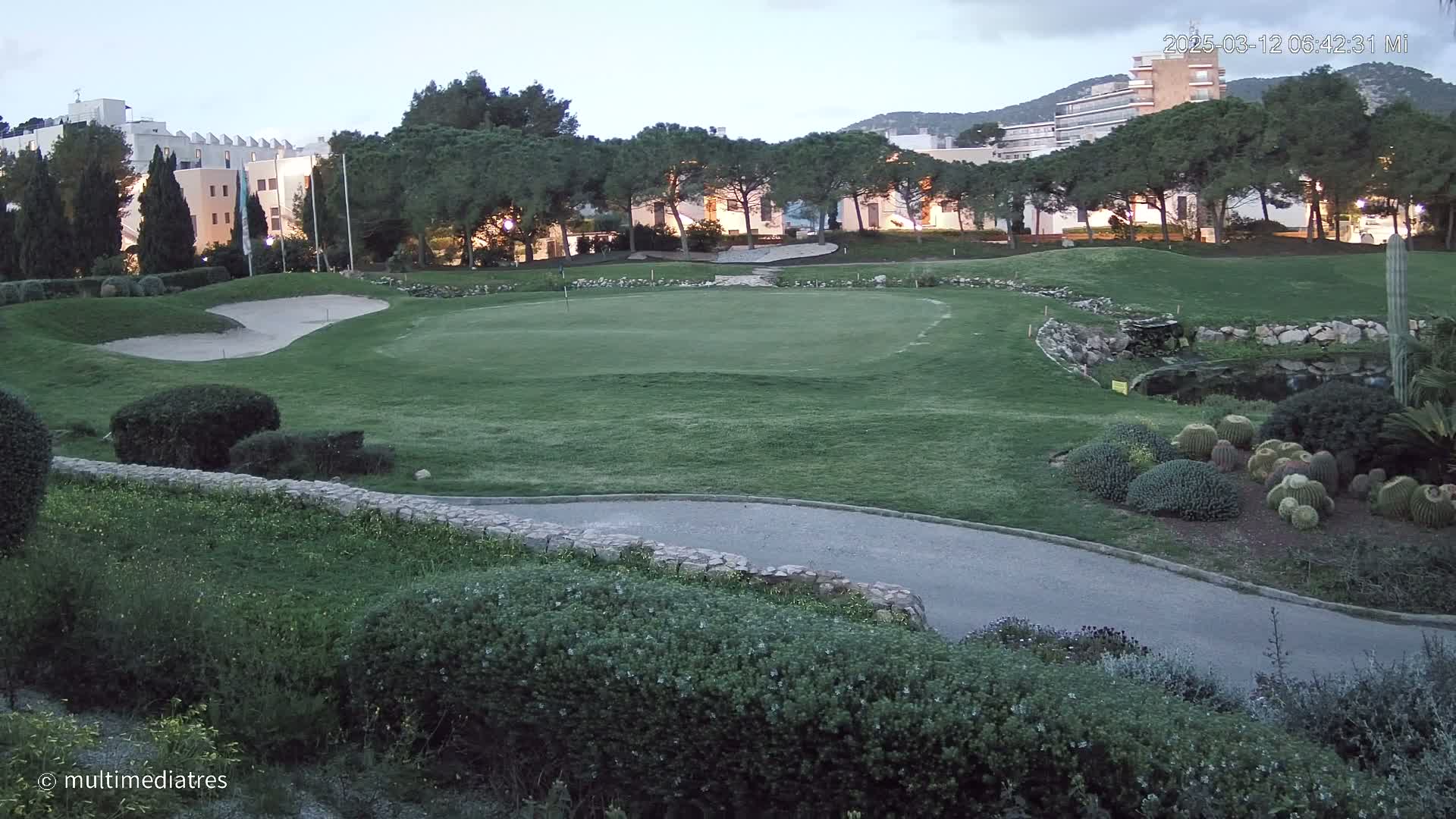 A golf course green and sand trap are visible, nestled amongst low shrubs and trees under a twilight sky near buildings.