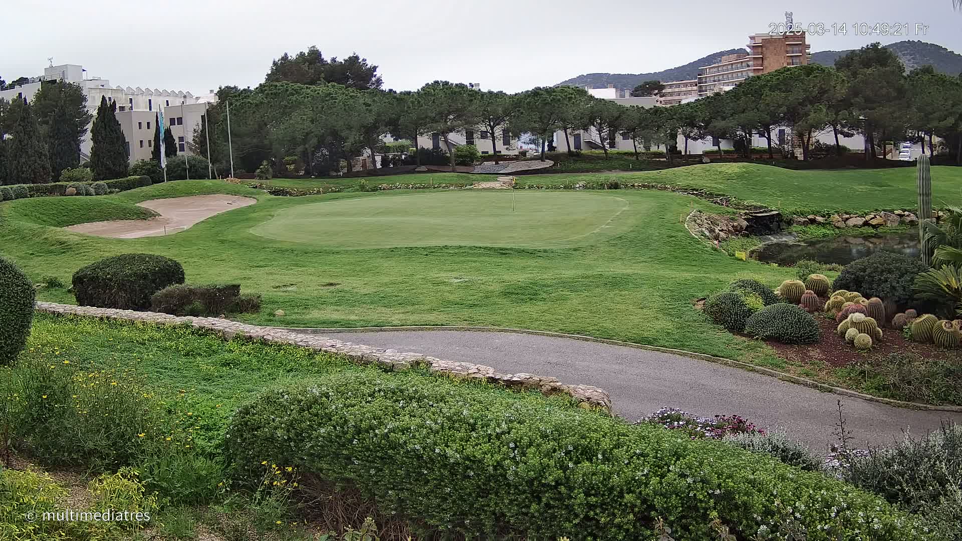 A golf course green, sand trap, and surrounding landscaping are visible under an overcast sky, with buildings and trees in the background.